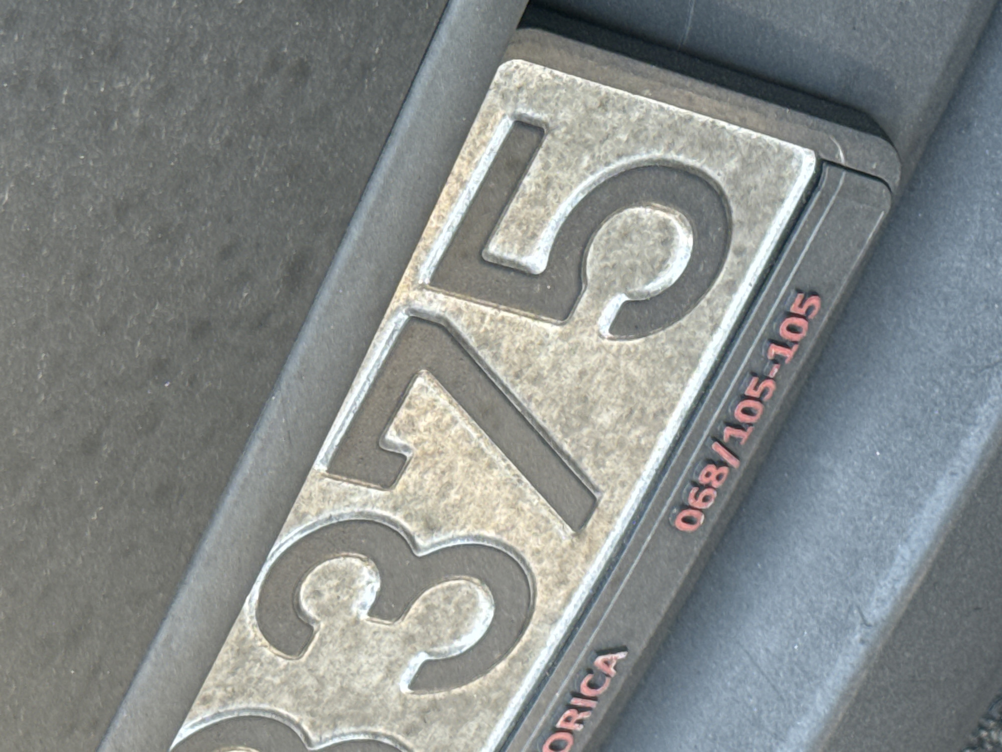 A close-up, eye-level shot of a license plate on the rear of a vehicle, seen from the side. The license plate is metallic grey with embossed red numbers and text. The visible portion of the license plate shows the numbers "375" prominently, with partial numbers to the left, and a series of numbers "068/105-105" in red text on the lower section. The text "ORICA" is also partially visible in red to the left of the lower numerical sequence. The license plate is set into a black frame, which is attached to a dark grey textured surface, likely the bumper of the vehicle. The background is a dark, out-of-focus grey surface. The lighting is even, with no harsh shadows. The image does not provide any contextual information about the city of Trap, Montenegro, or any other environmental details.