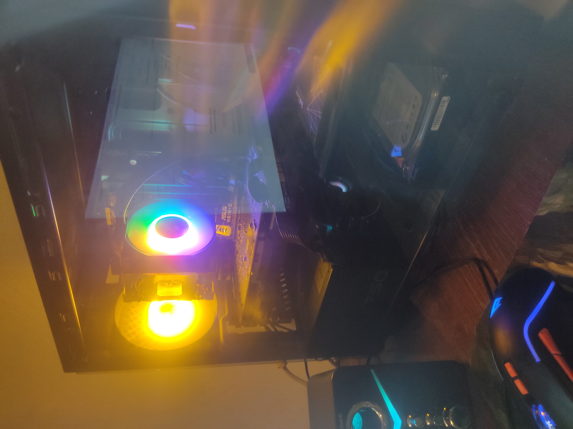 This is a close-up, angled shot of the interior of a computer tower, likely on a desk. The computer is illuminated by internal RGB lighting, with a prominent fan showing a vibrant rainbow spectrum of colors at the top, and another fan below glowing a bright yellow. The lighting creates a colorful and somewhat blurry effect on the components within the case.
Visible components include a hard drive mounted in the upper right section of the case. Below it, a motherboard is partially visible, with the text "ASUS H61M-D" clearly legible on a component. A power supply unit with the "QS 750" designation is also visible towards the right.
In the foreground, to the right of the computer tower, a gaming mouse with blue and orange LED accents is visible. To the left and below the computer, a speaker with blue and green lighting is also present. The overall scene suggests a gaming or computer enthusiast's setup. The lighting indicates it's likely nighttime or a dimly lit room. The presence of specific computer hardware components and peripherals are the main subjects. There are no people or animals visible. The background appears to be a wooden surface, possibly a desk. There are no overt environmental cues like weather or specific location identifiers, beyond the "Location context" provided, to suggest anything outside the immediate computer setup.
