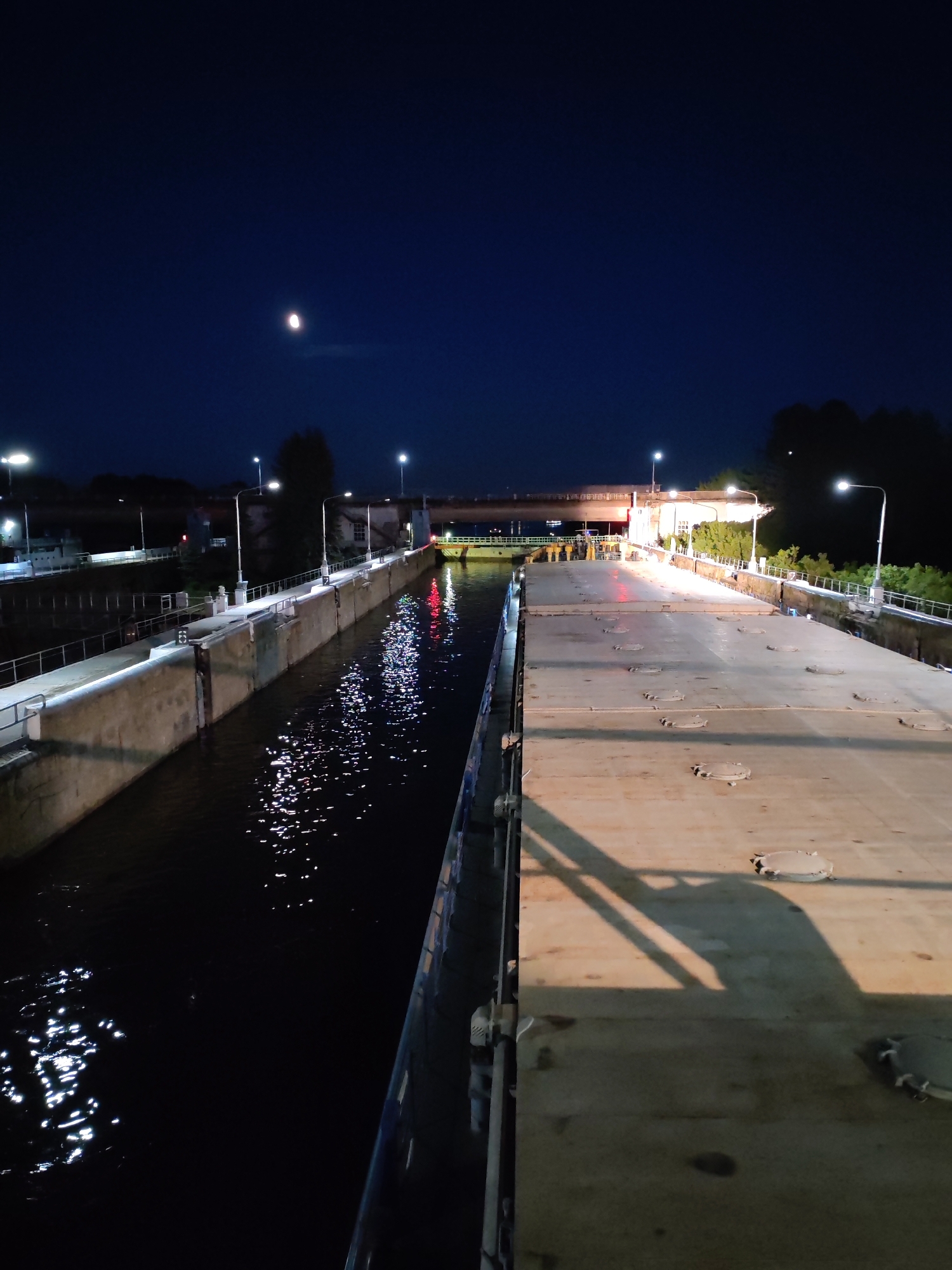 This is a night-time, vertical, medium shot of a canal lock in Bol'shie Mkhi, Russia. The sky is dark blue with a crescent moon visible in the upper left quadrant. Several streetlights illuminate the scene, casting light on the concrete walls and a large, flat-topped barge positioned in the right side of the lock. The water in the lock is dark, reflecting the artificial lights and the moon, creating a shimmering effect on its surface. In the background, a bridge spans the canal, with more lights visible on its structure and on the surrounding buildings. Trees can be seen in the distance on the right bank. The overall impression is one of a functional industrial area at night, with a serene yet somewhat stark atmosphere. There are no people visible in the image.