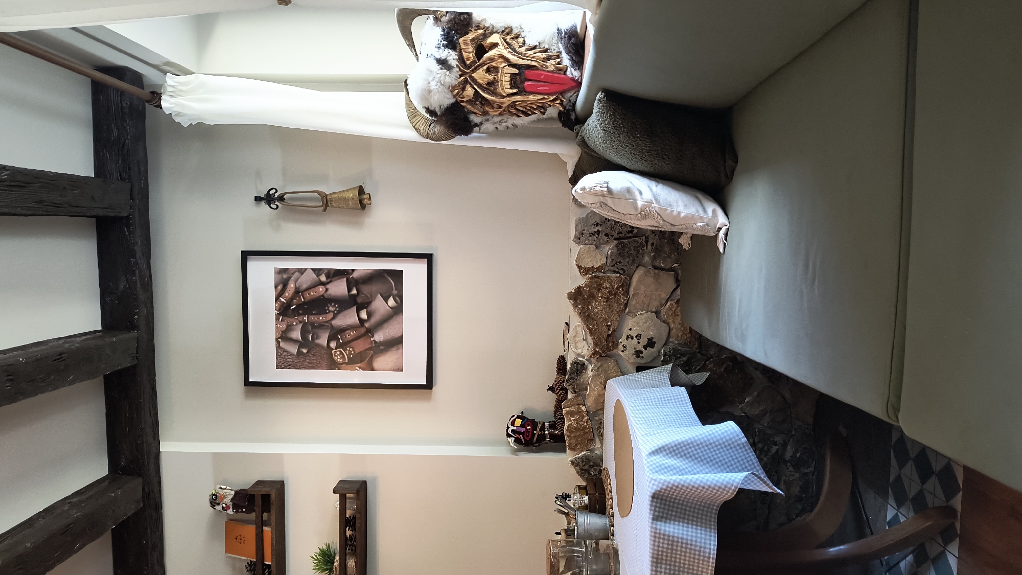 The image presents a room with an eclectic mix of decor elements. A sofa occupies a significant portion, draped with cushions and a decorative mask featuring horns and a carved wooden face. The room is adorned with a framed picture of cowbells and a vintage-style horn or bugle mounted on the wall. There's a stone wall section, juxtaposed with a blue-and-white checkered tablecloth on a table or surface below. A dark wooden beam or architectural element is visible on the left side. The overall ambiance suggests a rustic or traditional style, possibly within a residential or commercial space. The image provides no direct evidence to confirm that the scene is in Sofia, Bulgaria.