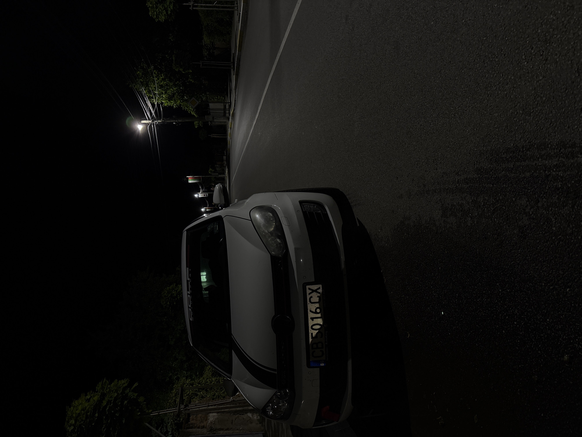The image depicts a white car parked on a street at night in Dolna Banya, Bulgaria. The car, which appears to be a Volkswagen Golf, has a distinctive black stripe running down the center of its hood and front bumper. A sticker partially visible on the top of the windshield reads "GOLF MOTORSPORT." The front license plate displays "CB 5016 CX," with the country code "BG" for Bulgaria, and a small "B" symbol, likely indicating the registration district.
The vehicle is positioned alongside an asphalt road that appears damp or wet, reflecting the dim light available. A white solid line marks the edge of the road. In the background, a solitary, brightly lit street lamp illuminates a portion of the scene, casting light on some trees and foliage that line the left side of the street. Numerous power lines extend from the pole holding the street lamp, silhouetted against the dark night sky. No activity or people are visible; the scene focuses on the stationary car in a quiet, nocturnal setting.