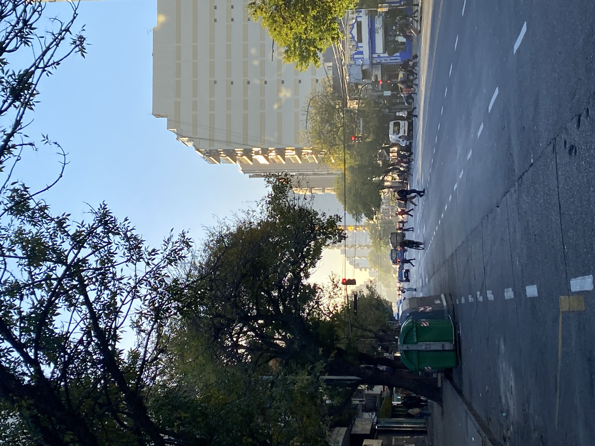 The image depicts a street scene in Buenos Aires, Argentina. A tall, light-colored building dominates the left side of the frame, partially obscured by a tree with dense foliage in the foreground. The street stretches into the distance, with several people visible crossing the road. Cars and a white van are also present. A green trash container sits on the sidewalk to the right. A traffic light is visible further down the street. The sky is pale blue and sunlight appears to be diffused.