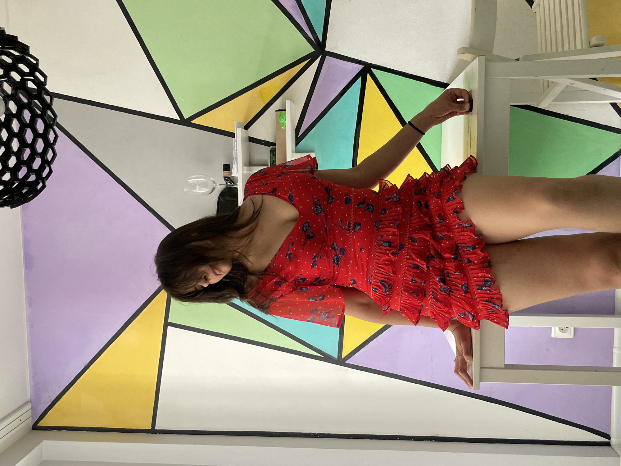 A woman in a red, polka-dotted dress with blue floral patterns and ruffled details is the sole subject of this image. She is leaning against a white table and chair set, with her body angled towards the right of the frame. Her right arm is extended, and her hand is resting on the edge of the table. Her face is partially obscured by her dark hair, and her eyes are downcast, suggesting a moment of reflection or relaxation.
The background is a striking geometric mural painted on a wall, featuring large, interlocking triangles and trapezoids in various pastel colors, including purple, yellow, green, and white, outlined in black. To the left of the woman, a black, hexagonal lamp hangs from the ceiling. A wine glass and a bottle are visible on a shelf behind her.
The lighting appears to be natural, likely from an indoor source or diffused daylight, casting soft shadows and highlighting the vibrant colors of the mural. The setting seems to be a contemporary indoor space, possibly a cafe, studio, or a home with an artistic design. There are no visible indications of the time of day or weather conditions. The overall mood is casual and serene. No text is visible in the image. The location context is Izhevsk, Russia.