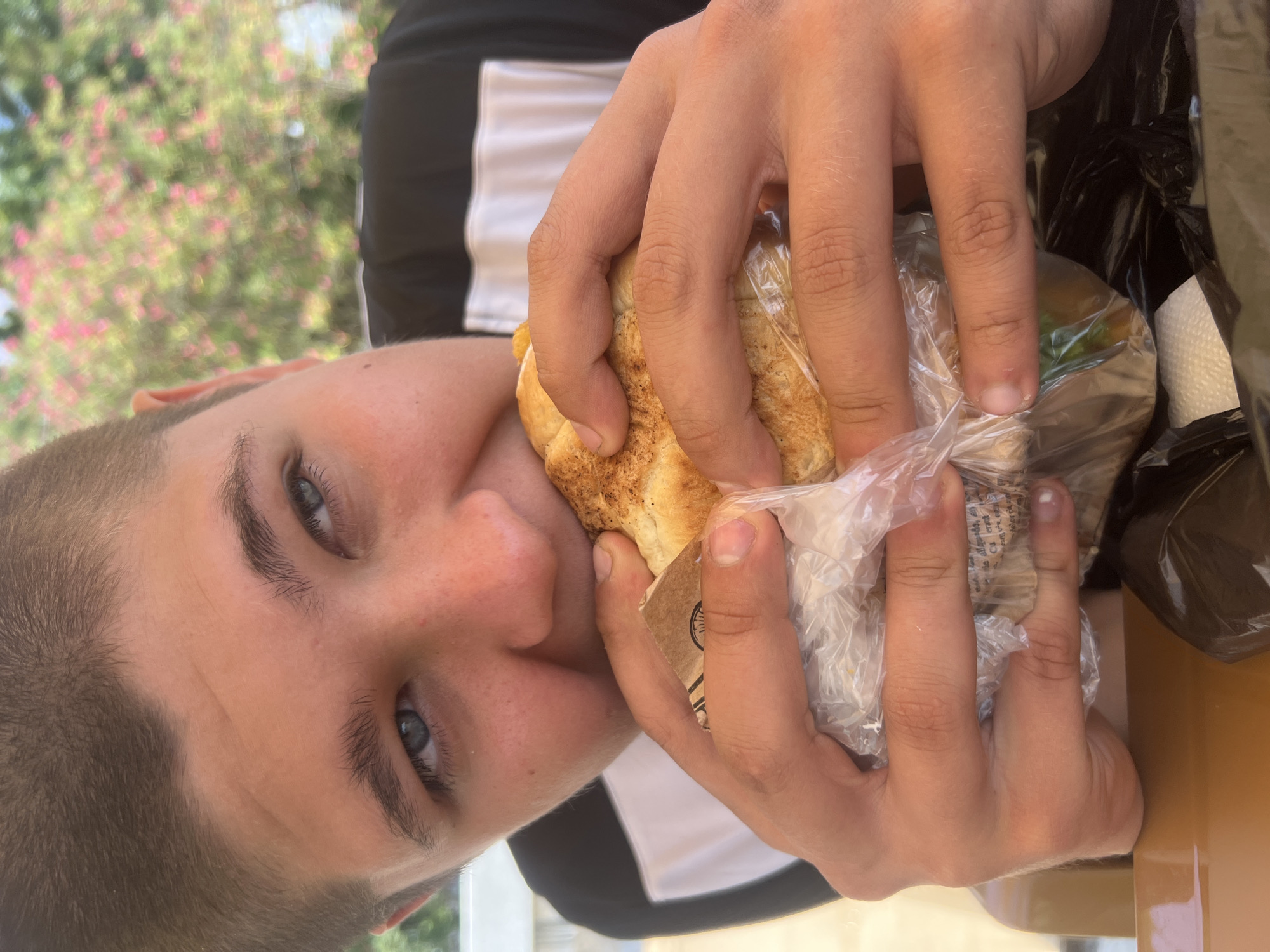 A close-up, eye-level, outdoor shot shows a young person with short brown hair biting into a sandwich. The person's head is tilted to their left, and their blue eyes are looking upwards and to their left. They are wearing a black t-shirt with a white stripe. The sandwich is wrapped in clear plastic and a piece of brown paper. The background is blurred with green foliage and pink flowers, suggesting a natural or garden setting, possibly in a cafe or restaurant. A brown table surface is visible in the foreground. The image does not contain enough information to determine the specific location within the city of Sokhumi, Abkhazia.