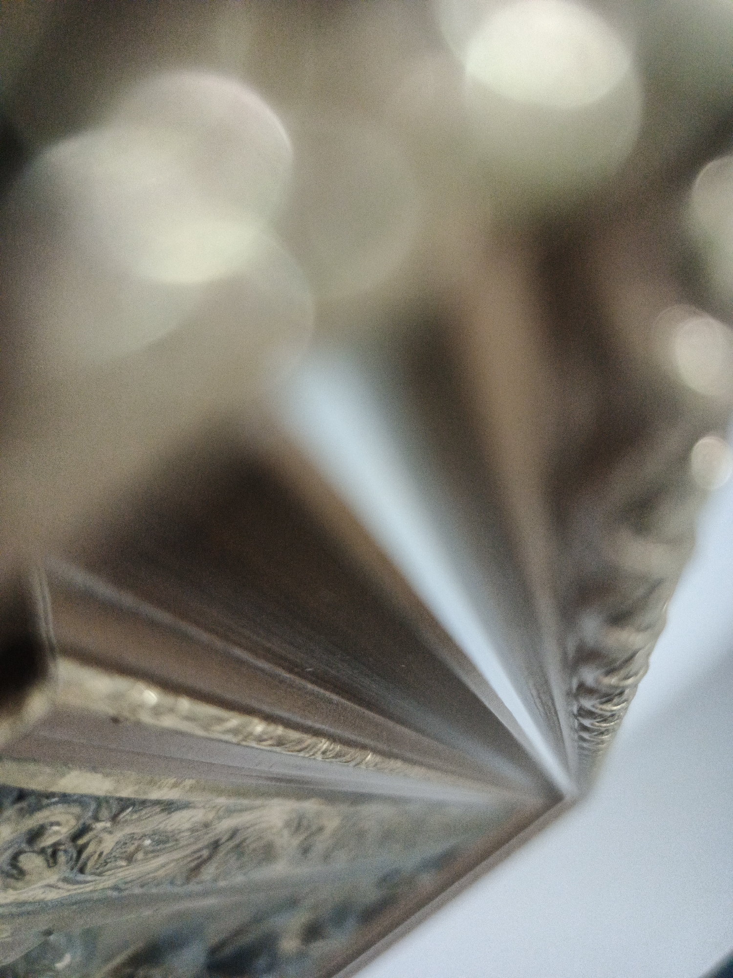 The image depicts a close-up, abstract view of what appears to be the corner of a picture frame with multiple layers or segments. The frame has a metallic, possibly golden or bronze, finish. The texture on the bottom left portion of the frame is rough and patterned. There are out-of-focus, light-colored orbs, likely bokeh, in the background, suggesting light sources behind the frame. The right side of the frame is adjacent to a white surface. The image provides no information about the city of Los Robles, Venezuela.
