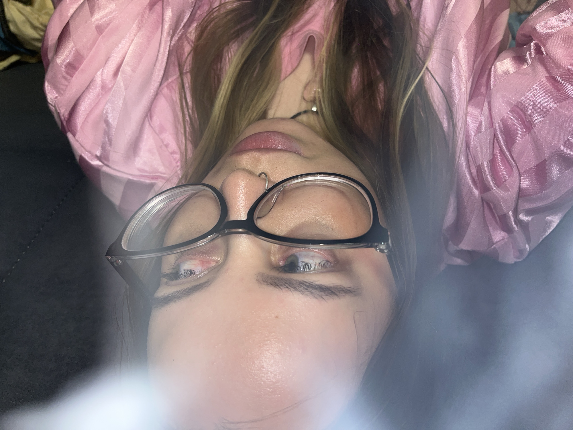 The image shows a close-up, distorted shot of a young person wearing glasses and a pink silky garment. Their face is partially obscured by the glasses and the angle of the photograph. The background appears to be a dark, possibly upholstered surface. The lighting in the image is uneven, with a bright glare obscuring a portion of the left side. The person's hair is visible, appearing to be medium-length and light brown with blonde highlights. The setting does not provide any discernible information about the city of Novopokrovskaya, Russia, or any specific location within it.