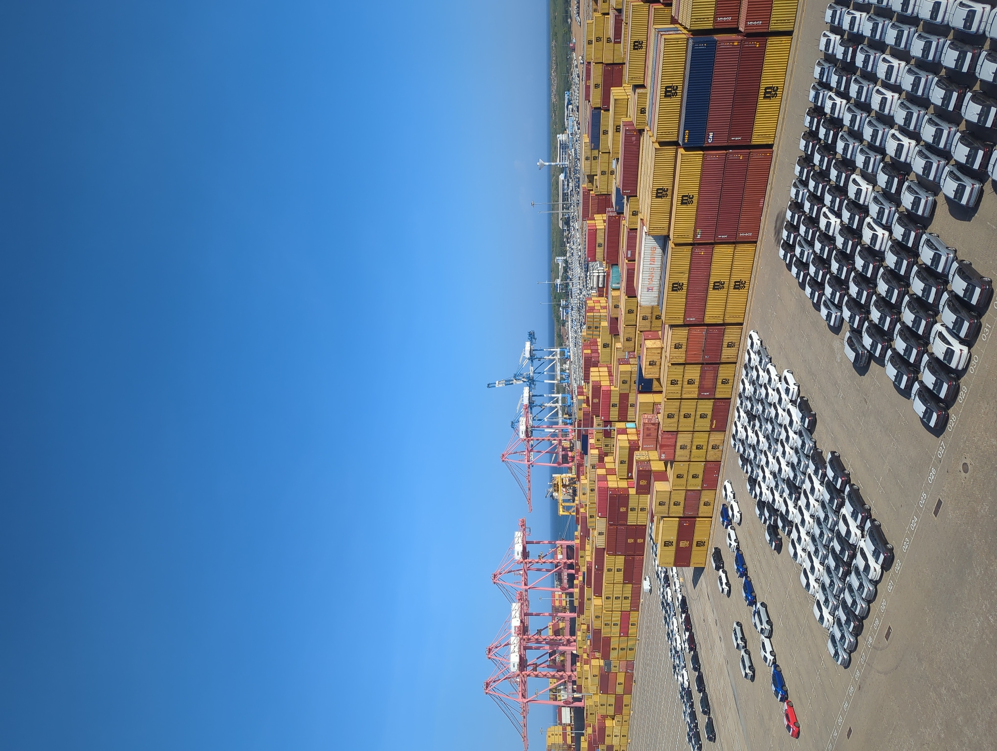 The image shows a large container terminal at the Port of Hambantota, Sri Lanka. The scene is dominated by stacks of colorful shipping containers, predominantly red and yellow, with some blue ones visible. Several large cranes, painted pink and blue, are positioned at the port, suggesting active cargo operations. In the foreground, a vast expanse of a paved surface is covered with rows of parked cars, mostly white and silver, arranged in an organized manner. The sky above is a clear, bright blue. The environment appears to be a sunny day in a busy port city.