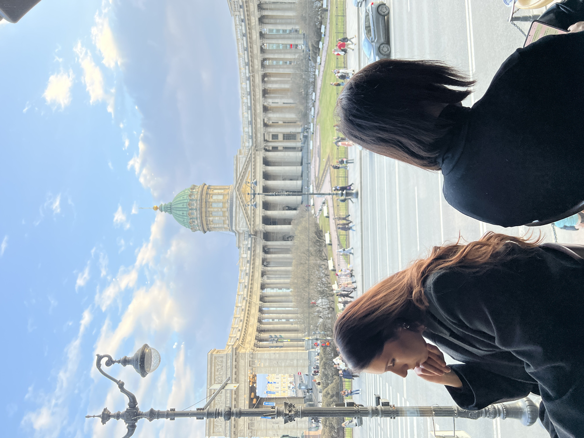 The image shows a view of Kazan Cathedral in Saint Petersburg, Russia, seen through a window from an elevated perspective. Two people are partially visible in the foreground, likely standing inside a building looking out at the cathedral. One has short, dark hair, and the other has long, brown hair. Both are wearing dark clothing. The cathedral is prominent in the background, with its distinctive dome and colonnade. A street runs between the cathedral and the viewer, with pedestrians and cars visible. The sky is blue with some clouds. A lamppost is also visible in the foreground. The photo appears to be taken from inside a building, looking out through a window.