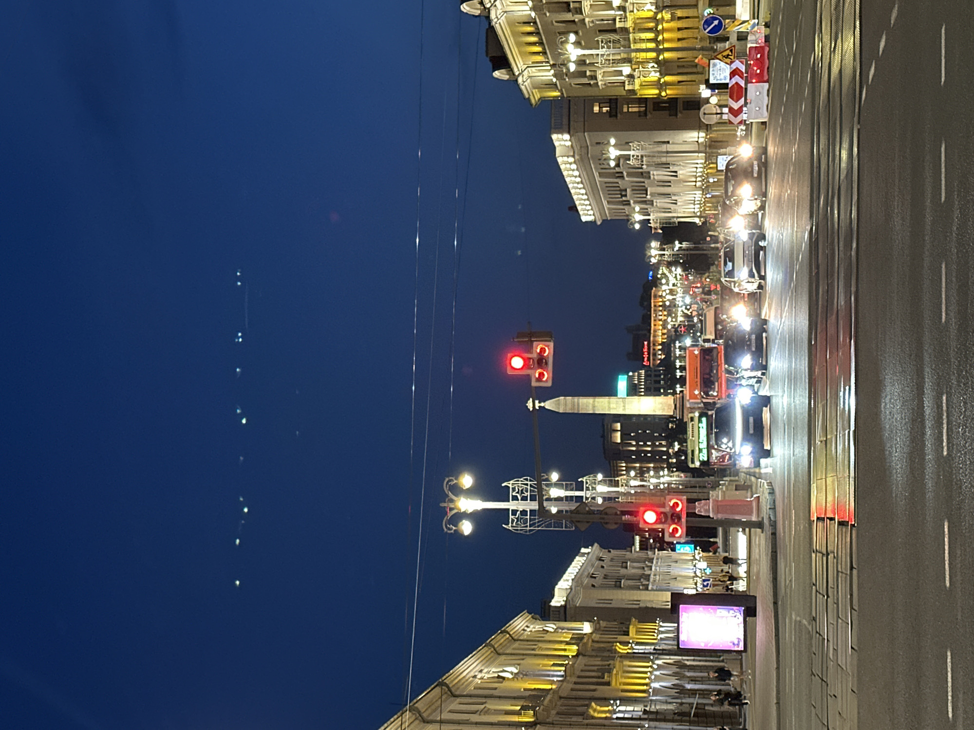The image captures an evening scene in Minsk, Belarus, specifically along Independence Avenue, near the iconic "Minsk Gates" or "Pillar Gate" at the entrance to the city from the west. The sky is a deep twilight blue, with a few faint airplane lights visible in the distance.
On the left side of the frame, a grand, illuminated building lines the avenue, its windows and architectural details lit with warm yellow light. A digital billboard displays a bright, abstract image. Pedestrians are visible walking on the sidewalk.
Dominating the center of the image is a tall, elaborate street lamp with multiple globes, also illuminated. Further down the avenue, a prominent obelisk, part of the "Minsk Gates" landmark, stands against the darkening sky. Traffic lights show a red signal.
The avenue itself is busy with vehicles. Several cars and a truck are visible, their headlights cutting through the dim light. The wet road surface reflects the lights of the traffic signals and the vehicles. On the right side of the avenue, another large, well-lit building mirrors the architecture of the one on the left. Traffic barriers and road signs are also present, indicating ongoing road work or traffic management. The overall atmosphere is that of a bustling urban environment transitioning from day to night.