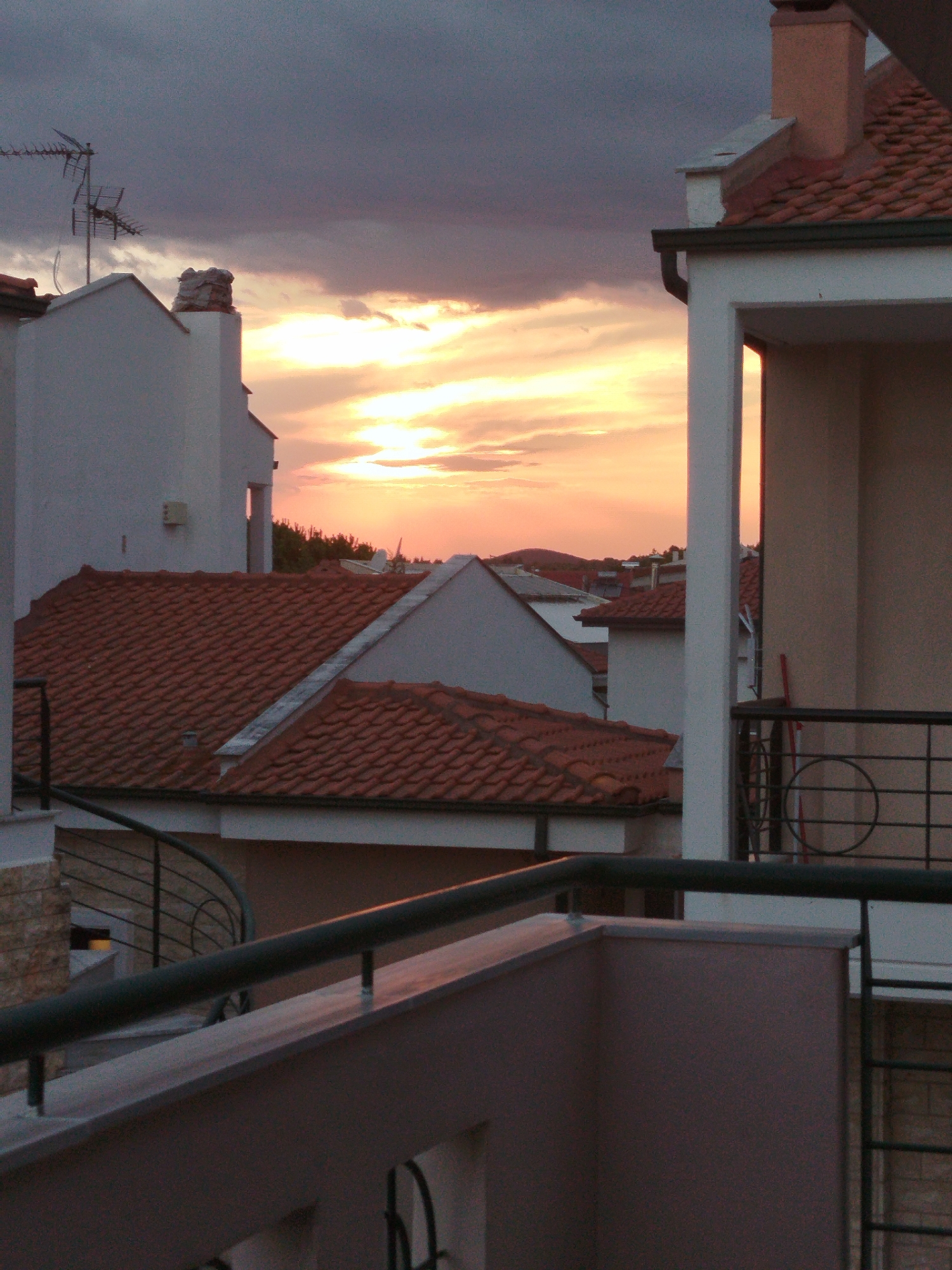 The image depicts a twilight scene of rooftops in a Greek town, likely Sithonia Municipal Unit, Greece, given the architectural style and the presence of terracotta tiled roofs. In the foreground, a balcony railing, painted dark green, extends horizontally across the frame. Behind it, the edge of another balcony is visible, with a light brown painted wall.
The middle ground is dominated by red-tiled roofs of various buildings. Some have chimneys, including one with a distinctive, rough-hewn stone top. The sky above is a dramatic mix of dark gray clouds and bright orange and yellow streaks of sunset light, indicating the sun is low on the horizon. A television antenna is visible on top of one of the white buildings.
In the background, more buildings with tiled roofs and some distant hills can be seen, partially obscured by trees. The overall impression is of a quiet, residential area at the end of the day. There are no people visible in the image, and the scene is static. The lighting suggests it is either sunrise or sunset.