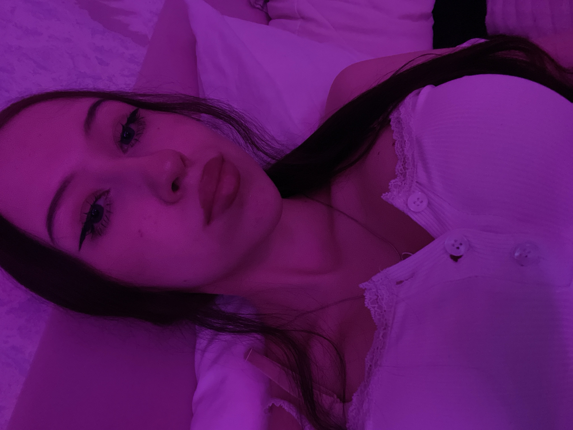 The image is a close-up selfie of a young woman with dark hair and dark eyes. She is wearing a white ribbed tank top with lace trim and buttons down the front. The lighting in the image is predominantly pink and purple, suggesting it might be from a light source like a colored LED or a party setting. Her makeup includes winged eyeliner and pink lipstick. She appears to be lying down, possibly in a bed, as a white pillow is visible in the background. The overall atmosphere is intimate and personal. There is no information in the image to indicate the location is Votkinsk, Russia.