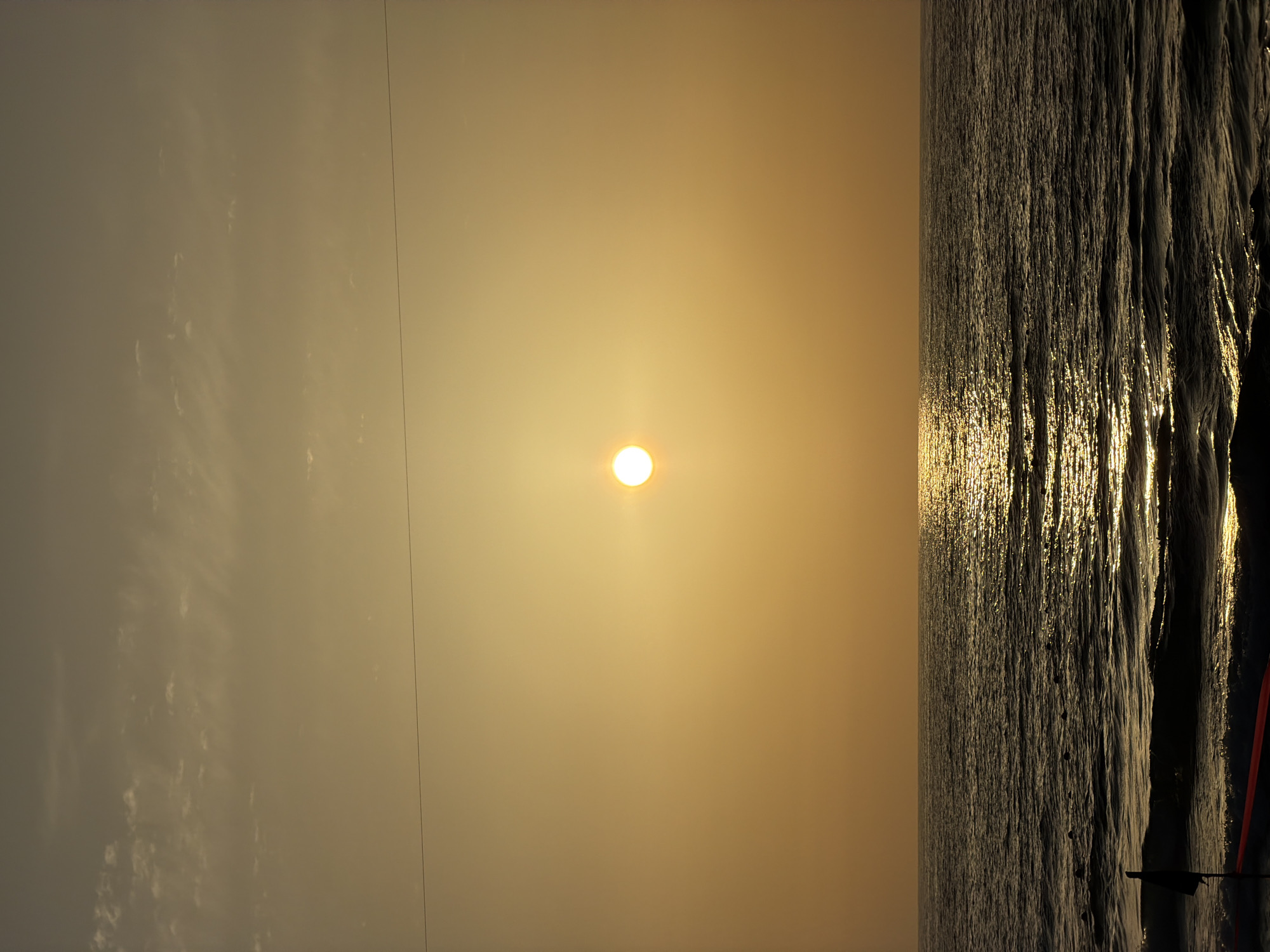The image depicts a sunset scene at Tel-Aviv beach, Israel. The sun is centered in the sky, casting a golden glow. The sea is on the right side, with light reflecting off the water. The sky on the left has a light cloud cover.