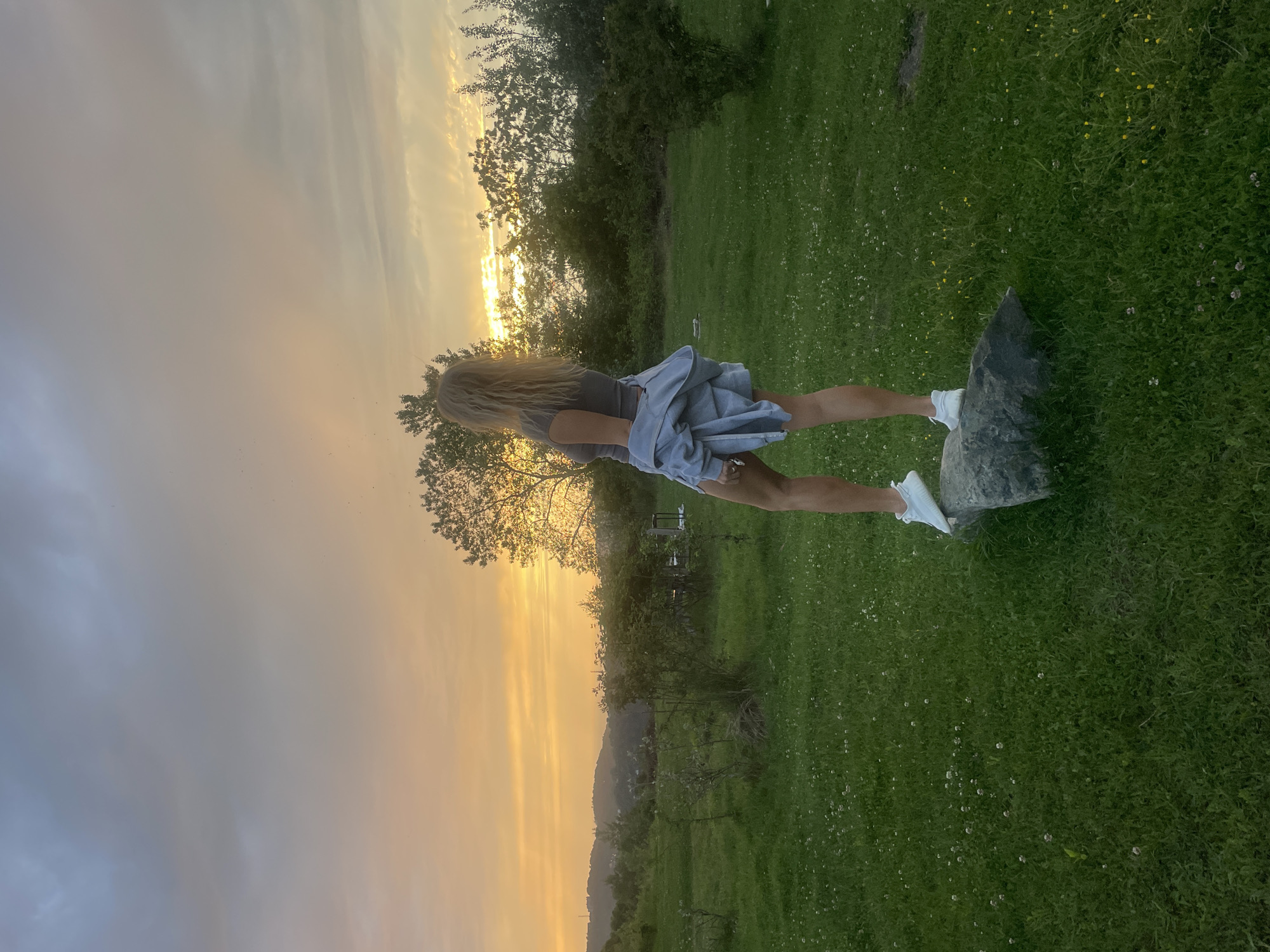 A woman with blonde hair stands on a rock in a grassy field, facing away from the camera and towards a sunset. She is wearing a grey shirt, a light blue jacket, and white shoes. The sky is a mix of yellow and grey clouds. Trees and hills are visible in the background. The location is Baranovka, Russia.