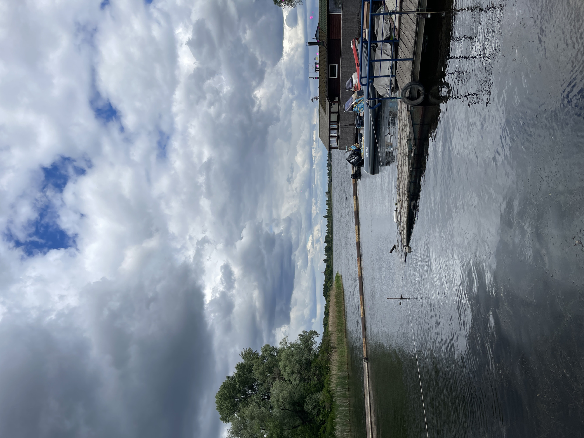 The image depicts a landscape near Vishen'ki, Ukraine. A body of water, likely a lake or river, dominates the scene. A wooden dock extends into the water with a small inflatable boat moored alongside it. The sky is filled with heavy, dark clouds, indicating a possibly inclement weather. Some green trees and grassy areas flank the edge of the water, and reeds border the waters edge. A wooden building stands at the edge of the dock. A single line extends into the water.