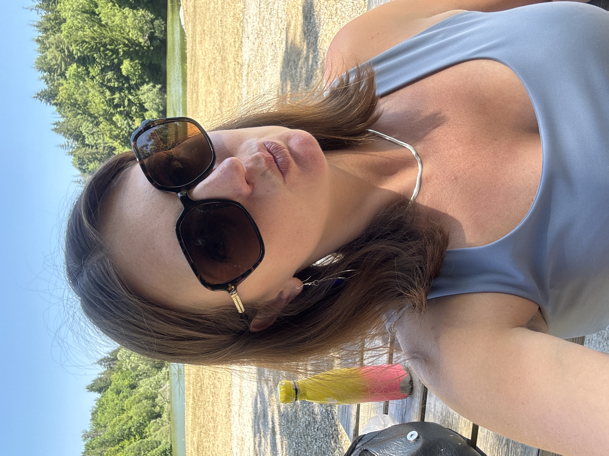 A woman is taking a selfie outdoors. She is wearing large, dark-rimmed sunglasses and a light blue tank top. She has brown hair and is wearing a thin silver necklace and long, dangling earrings. Her lips are pursed. She is sitting at a wooden picnic table, and a brightly colored water bottle is visible on the table to her left. Behind her, there is a body of water, likely a lake, with a densely wooded hillside beyond it under a clear blue sky. The area appears to be a park or recreational area.