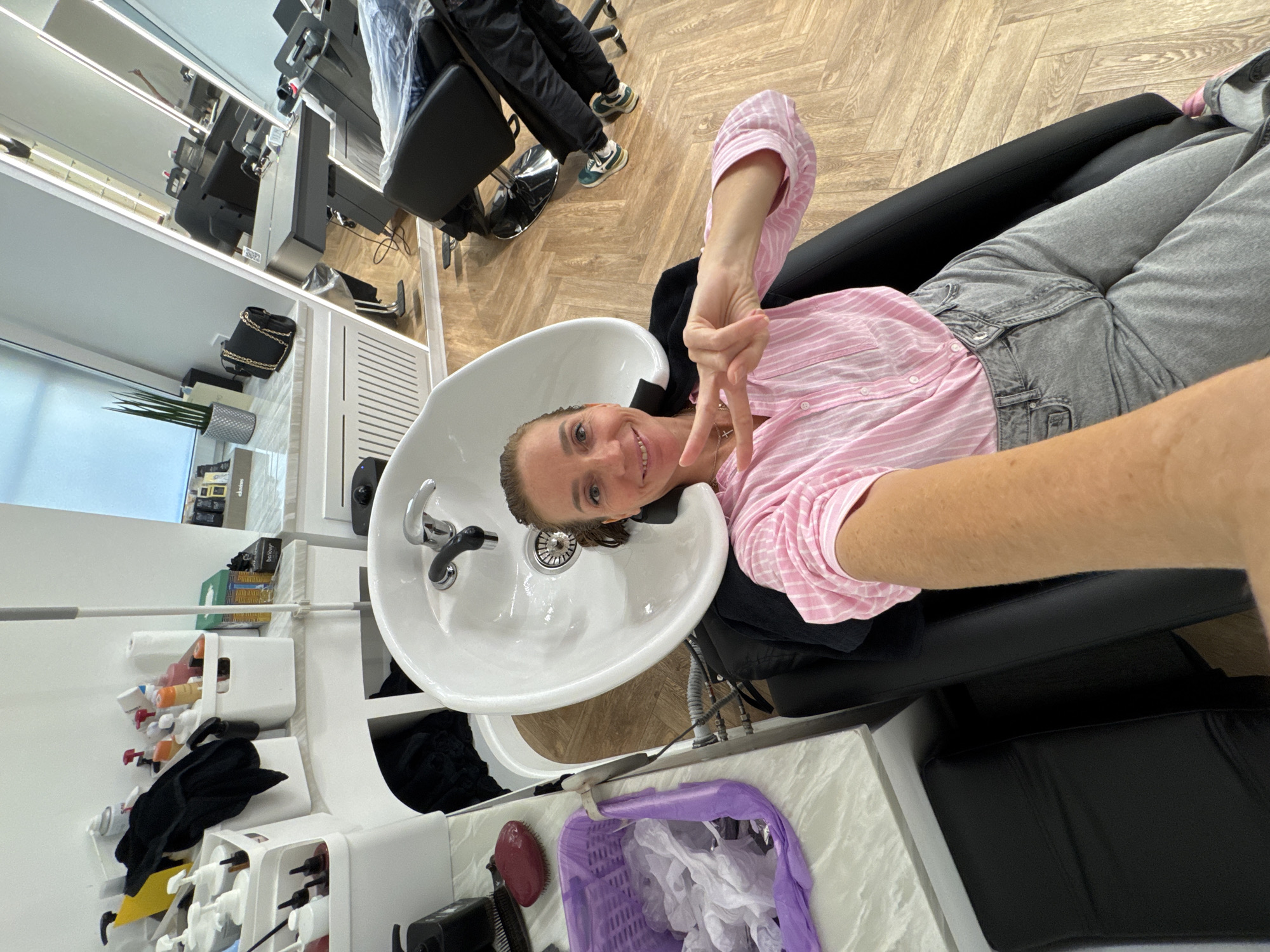 A woman with light-brown hair and a pink-and-white striped shirt lies in a salon chair with her head in a white shampoo sink, posing for a selfie with a peace sign. She is wearing grey jeans and a necklace. The salon has a modern interior with white walls and cabinets, a wooden floor, and black salon chairs. Several hair products and accessories are visible near the sink, along with a purple trash bin filled with white paper. The location is likely a hair salon, potentially in Saint Petersburg, Russia. Other salon chairs and styling stations are visible in the background.