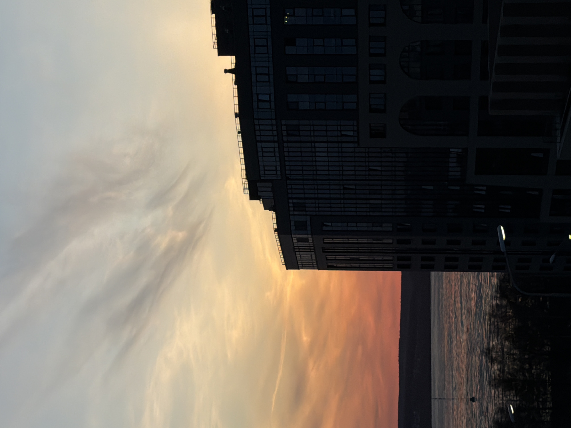 A horizontally oriented photograph depicts a high-rise building against a sky at sunset. The building, positioned on the right side of the image, has a dark facade with numerous windows and geometric patterns. To the left of the building, the sky is filled with hues of yellow and orange, indicating either sunrise or sunset. Below the sky, a body of water reflects the colors, with a dark shoreline visible in the lower section of the image. Street lights are visible near the water's edge. The image appears to have been rotated. The location is Izhevsk, Russia.