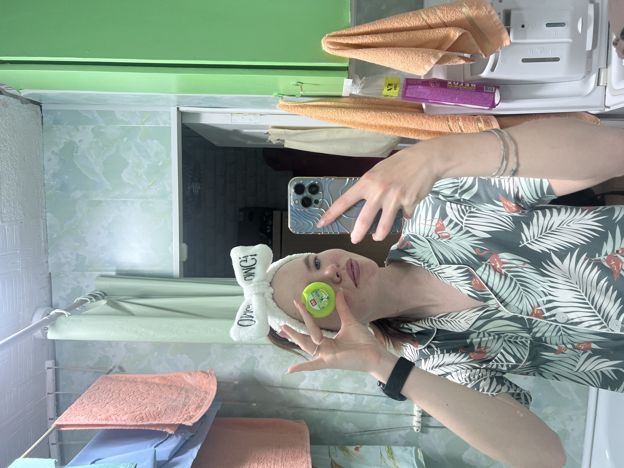 This image, reportedly captured in a bathroom in Chaikovsky, Russia, depicts a young adult female taking a mirror selfie. The subject, positioned centrally, is a fair-skinned individual with brown hair pulled back, wearing a white terrycloth headband with the reversed text "OWO C" (likely "OMG"). She is wearing a short-sleeved, collared pajama top featuring a dark green background with a white tropical leaf pattern and orange flamingo illustrations. Her left hand holds a silver-colored Apple iPhone, likely a Pro Max model due to its triple camera array and LiDAR sensor, encased in a clear cover with a blue, wavy, or ribbed design. Her right hand is extended towards her face, holding a small, round, bright green plastic container in front of her left eye, creating the appearance of an oversized, artificial eye. The container bears the brand "THAI," "FRUIT NATURE" text, and a white floral graphic, indicating it is a small balm or inhaler, weighing 2g. A silver ring is visible on her right ring finger. Her left wrist displays a black smartwatch with a fabric strap, and her right forearm shows two thin, braided silver bracelets. Her fingernails are manicured with a light polish. The bathroom environment features light green, possibly marble-effect wall paneling that shows some wear and tear, particularly at the seams and near the ceiling. The ceiling is white and textured. To the left of the subject, a white extendable clothes drying rack is mounted on the wall, holding several towels including a light blue and a peach-colored one. A light green shower curtain, hanging on a white rod with green rings, is pulled partially open behind the drying rack. To the right of the subject, a section of a green cabinet or shelving unit is visible. Two more peach-colored towels hang on green hooks on this unit. Below these towels and the green cabinet, a white front-loading washing machine is partially visible, showing its detergent dispenser drawer with markings like "MAX," "I," "II," and a flower symbol. A purple bottle, possibly a cleaning or laundry product with "DETOX" visible on its label, is contained within a clear plastic bag and is hanging or resting near the washing machine. A white door, slightly ajar, with visible silver hinges, is reflected in the mirror, and a beige or off-white fabric item hangs from it. The bottom left of the reflection shows the edge of a white bathtub