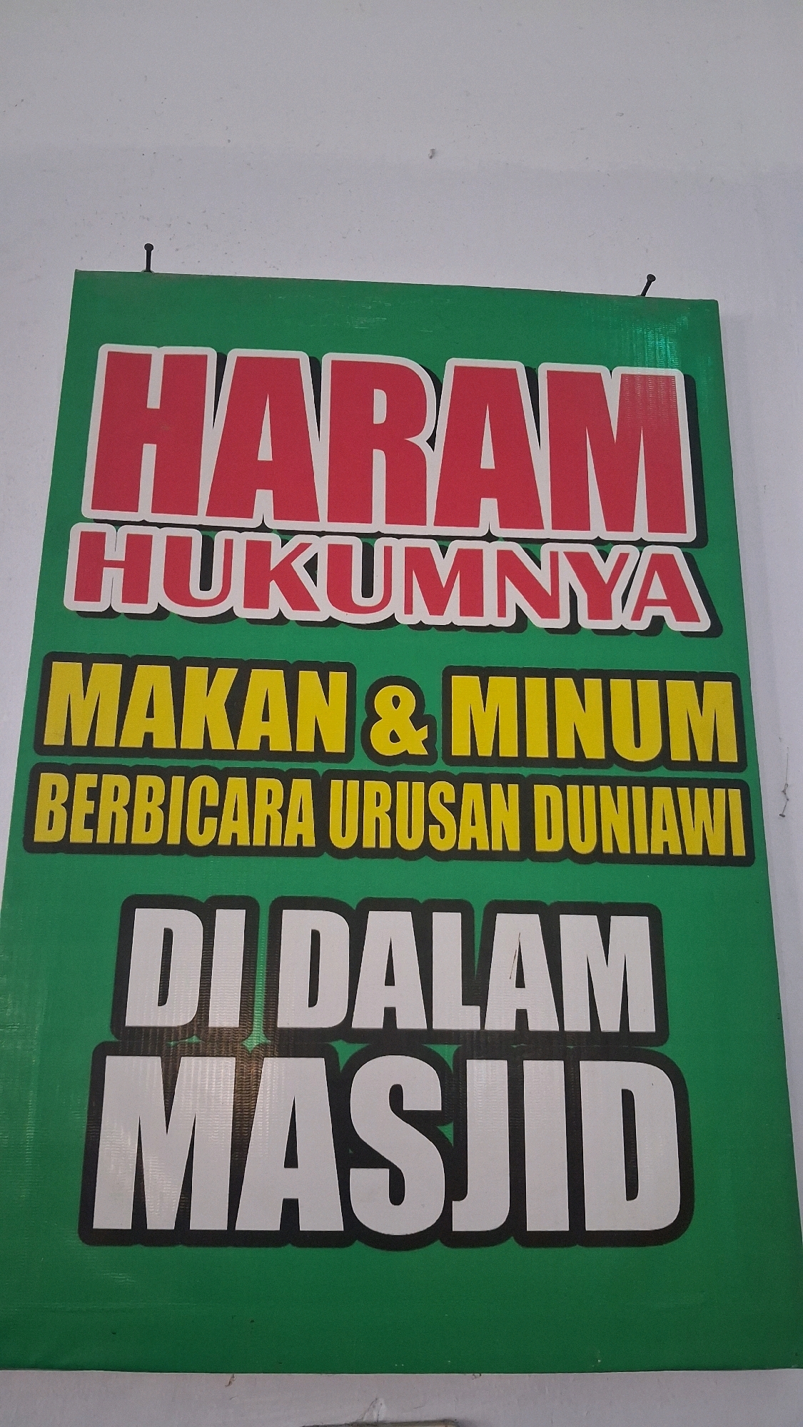 A green banner with white and red text is displayed on a white wall. The text states "HARAM HUKUMNYA MAKAN & MINUM BERBICARA URUSAN DUNIAWI DI DALAM MASJID". The banner is attached to the wall with a small nail. The banner appears to be located in front of a building. Based on the text, this banner is likely located in a mosque in Giriwarno, Indonesia, as it is a warning against eating, drinking, and engaging in worldly conversations within the mosque premises, which are considered forbidden (haram) in Islamic law.