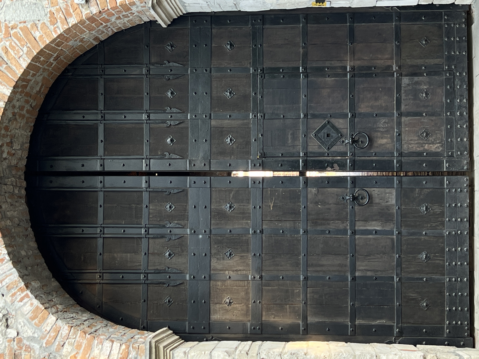 This image captures a large, imposing wooden gate, likely an entrance to a historic building or fortress in Zbarazh, Ukraine. The gate is constructed from dark, weathered wood panels, heavily reinforced with numerous black metal straps and rivets that create a grid-like pattern across its surface.
Each of the two main door panels features decorative ironwork, including diamond-shaped plates with raised centers and smaller, ornate, scrolled metal embellishments. A prominent metal ring handle is affixed to each door panel, set within a square escutcheon plate, indicating its function for opening and closing.
The gate is set within a robust arched entryway. On the left, a sturdy brick arch curves around the door, displaying detailed masonry. Lighter-colored stone elements are visible along the top and bottom of the frame, complementing the brickwork.
Bright light shines through a horizontal gap where the two door halves meet, and also along the bottom edge of the gate, suggesting it is daytime and there is an illuminated space beyond or directly behind the door. The overall impression is one of strength, history, and a secure passage. No people are visible, and there is no discernible text in the image.
