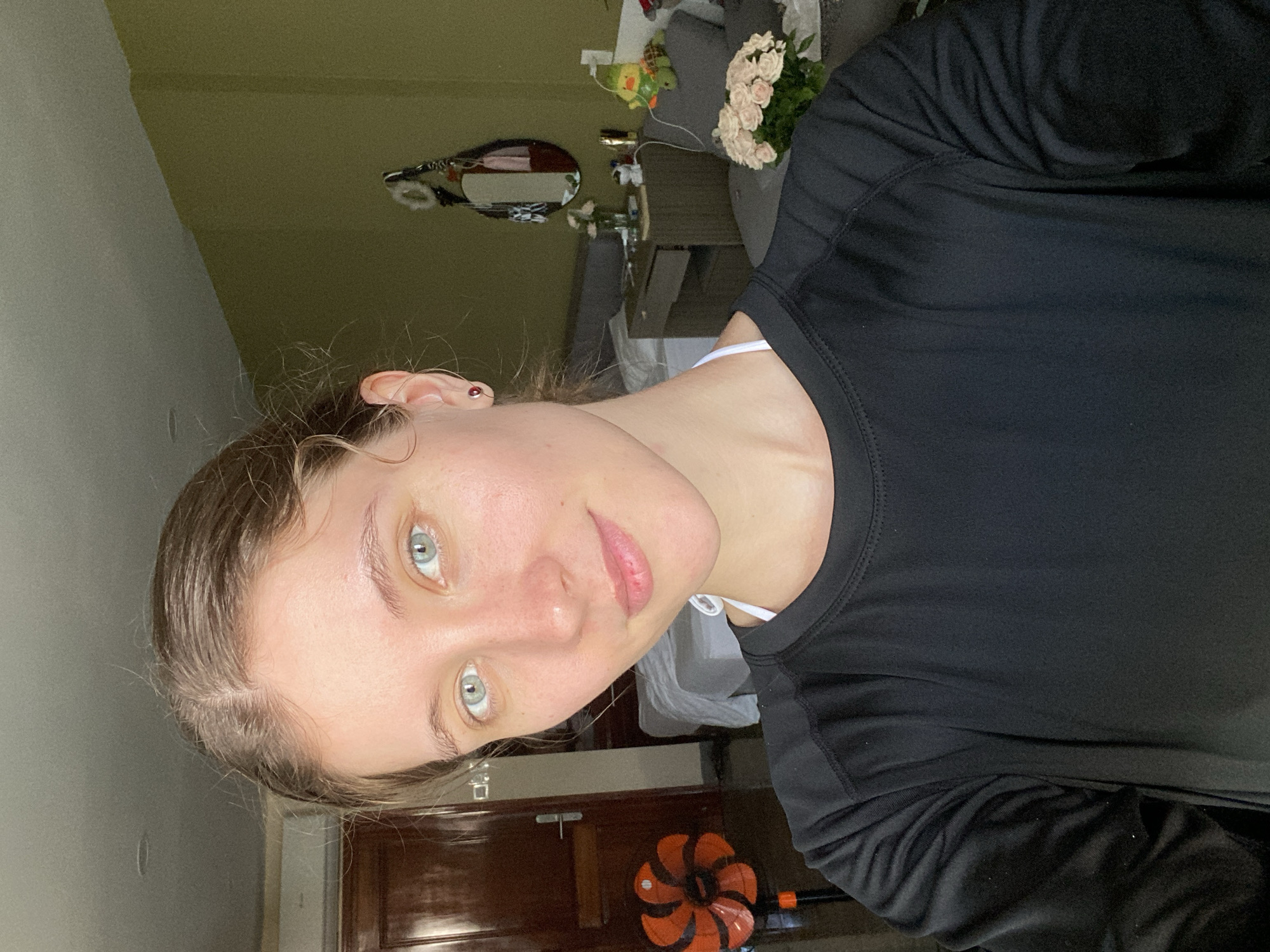 A young woman with light skin and blue eyes, her hair pulled back, poses for a selfie in what appears to be a bedroom in Bac Nha Trang Ward, Vietnam. She is wearing a black long-sleeved shirt with a white strap visible underneath. The background shows a doorway, a bed, a bedside table with some items on it, and a wall painted a muted green. A floor fan with orange blades is visible to the left of the woman.