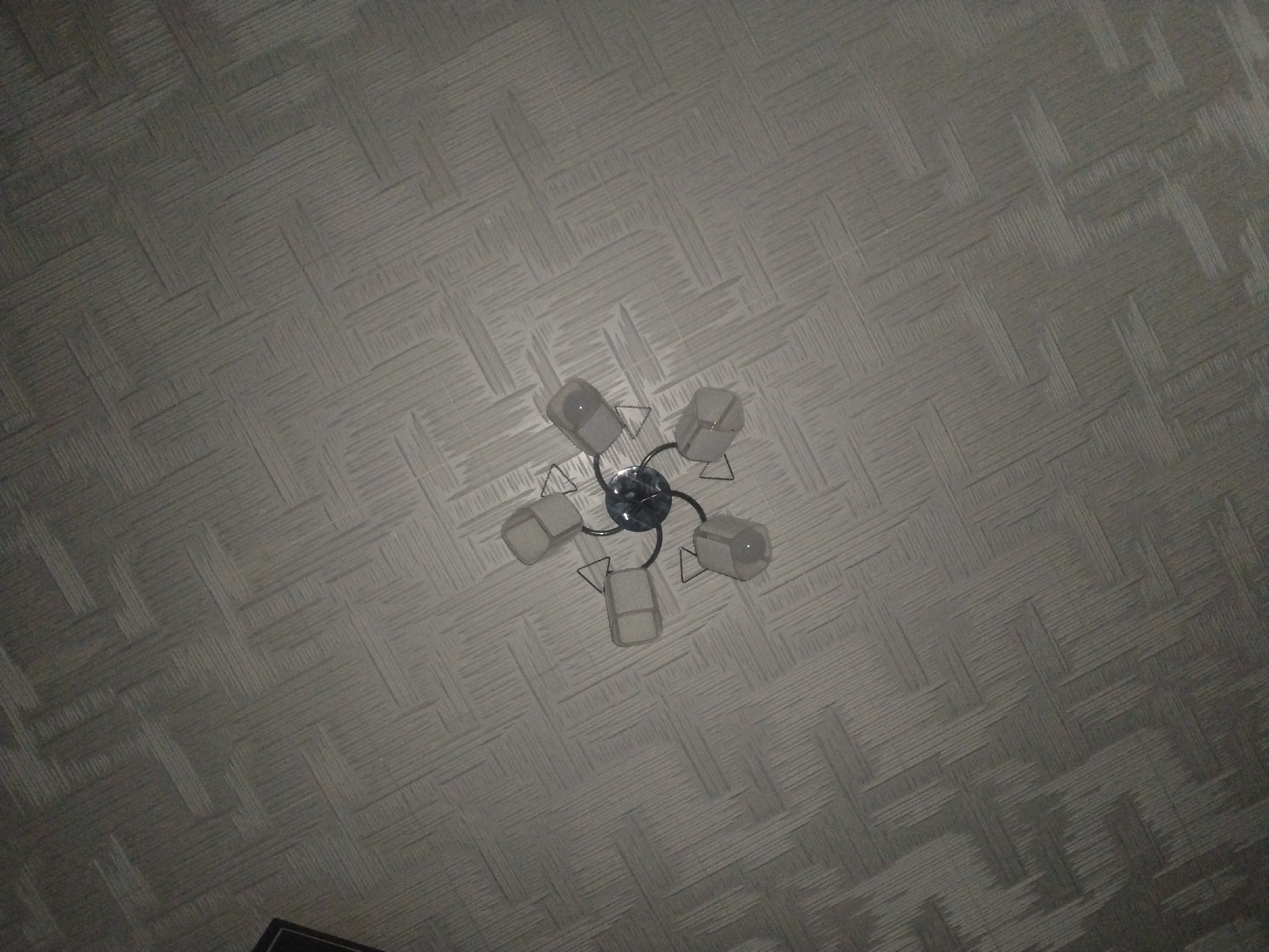 The image shows a chandelier with five lights attached to a textured ceiling. The chandelier has a circular base and arms that extend outwards, each holding a light fixture with a white, frosted glass shade. The ceiling has a repeating woven pattern in a light gray color. The image is taken from below, looking directly up at the ceiling. The location of the scene is in Zarafshan City, Uzbekistan.