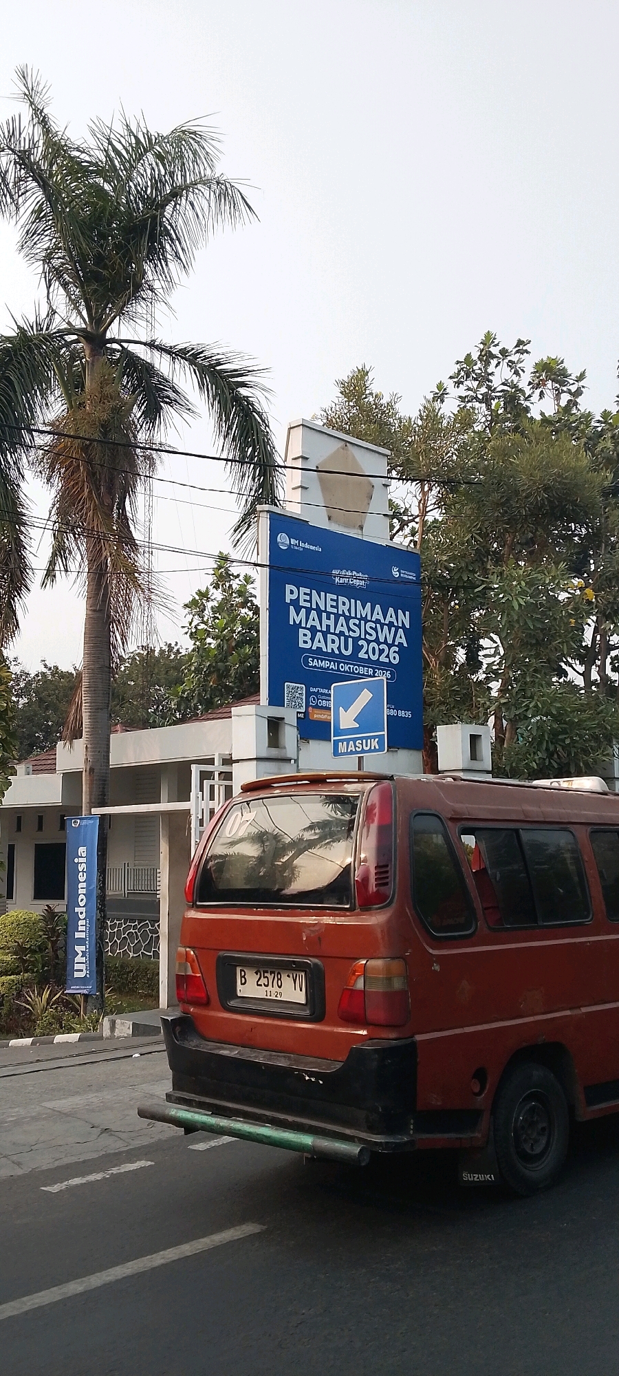 The image shows a street scene in Bekasi, Indonesia, featuring a street sign and a small orange van. The street sign, predominantly blue, displays text in Indonesian, including "PENERIMAAN MAHASISWA BARU 2026" (New Student Admissions 2026) and "SAMPAI OKTOBER 2026" (Until October 2026). Below this, a QR code and a phone number are visible, along with a directional sign indicating "MASUK" (Enter) with a downward arrow. The van is parked on the right side of the image and has a license plate that reads "B 2578 YV". Palm trees and other foliage are present in the background, suggesting a tropical or subtropical climate. The environment appears to be a relatively clear day.