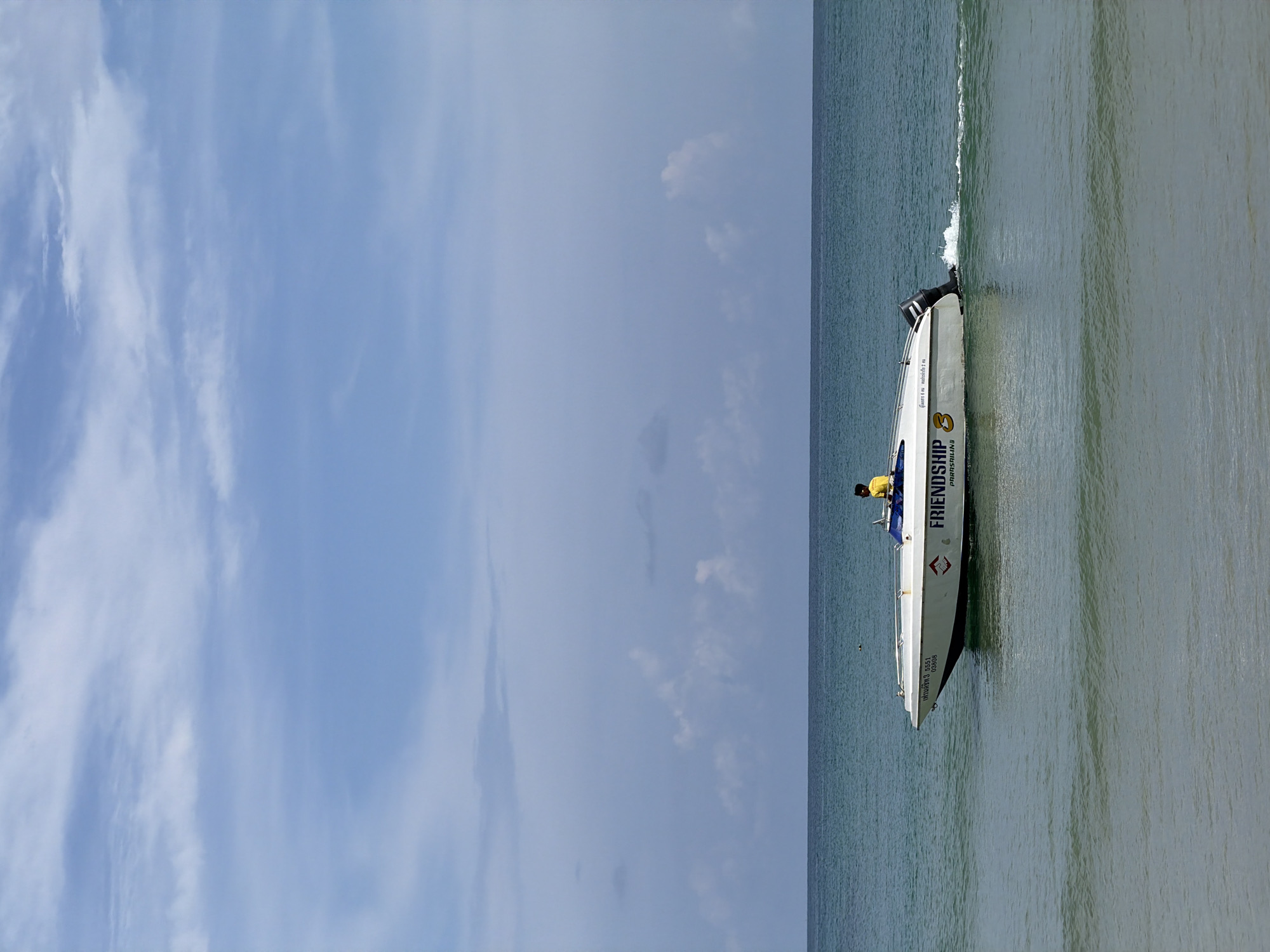 A white speed boat with "FRIENDSHIP" written on its side is cruising in the ocean under a partly cloudy sky. A person wearing a yellow shirt is seated in the boat, seemingly at the helm. The boat is leaving a wake behind it, indicating it is moving. The ocean is calm with a few gentle waves. The horizon is visible in the distance, separating the sea from the sky. The scene appears to be a scenic view of the ocean.