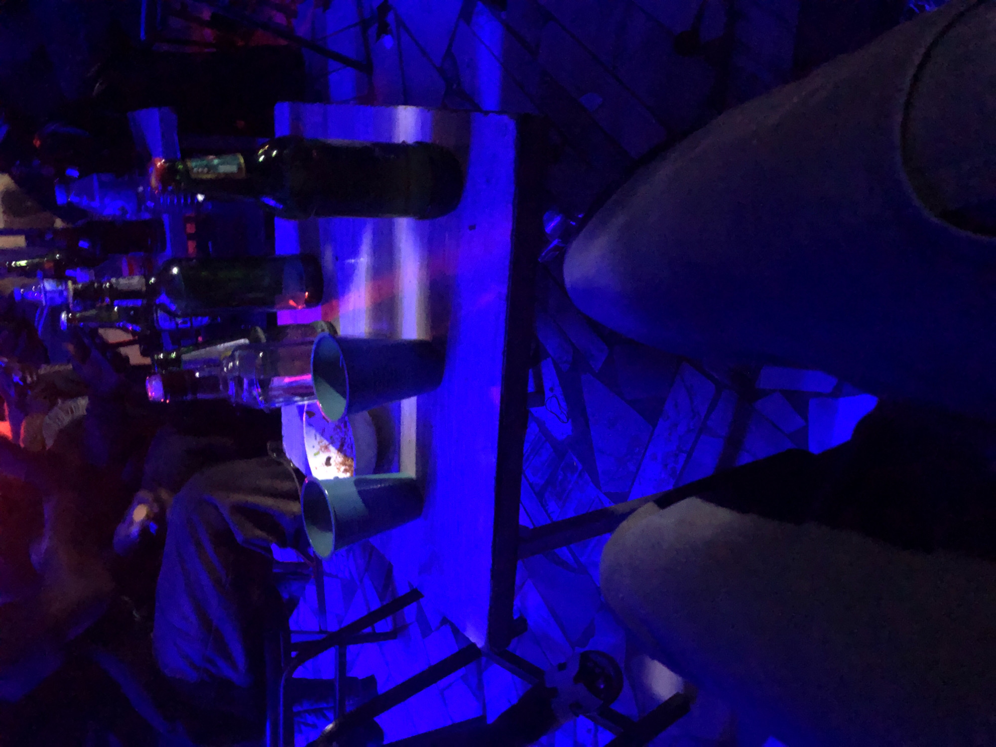 This image depicts an indoor scene, likely a bar or club, located in an unknown city and country. The perspective is from a seated individual, with their legs (clad in trousers or jeans) visible in the immediate foreground, positioned below a light-colored table.
On the table closest to the camera, illuminated by a strong blue light with hints of pink and red, are two light blue/teal plastic cups, a white bowl containing remnants of food (possibly rice or grains), and an empty brown glass bottle standing upright.
Further back, on an adjacent table, several more glass bottles are visible, primarily beer bottles of varying colors (green and clear), along with one clear plastic water bottle. These bottles are arranged in an informal manner.
In the background, multiple indistinct figures are present, mostly visible as shadowy outlines or partial forms due to the low and colored lighting. They appear to be seated at tables, suggesting a social gathering. Dark-framed chairs are visible supporting the tables.
The room's flooring consists of patterned tiles, visible under the tables and around the lower periphery of the image. The dominant lighting is a deep blue, casting strong shadows and highlighting surfaces with a cool hue, while occasional pink or red light sources add accents, particularly on the table surfaces and reflections on bottles. The overall atmosphere is dark and casual.
