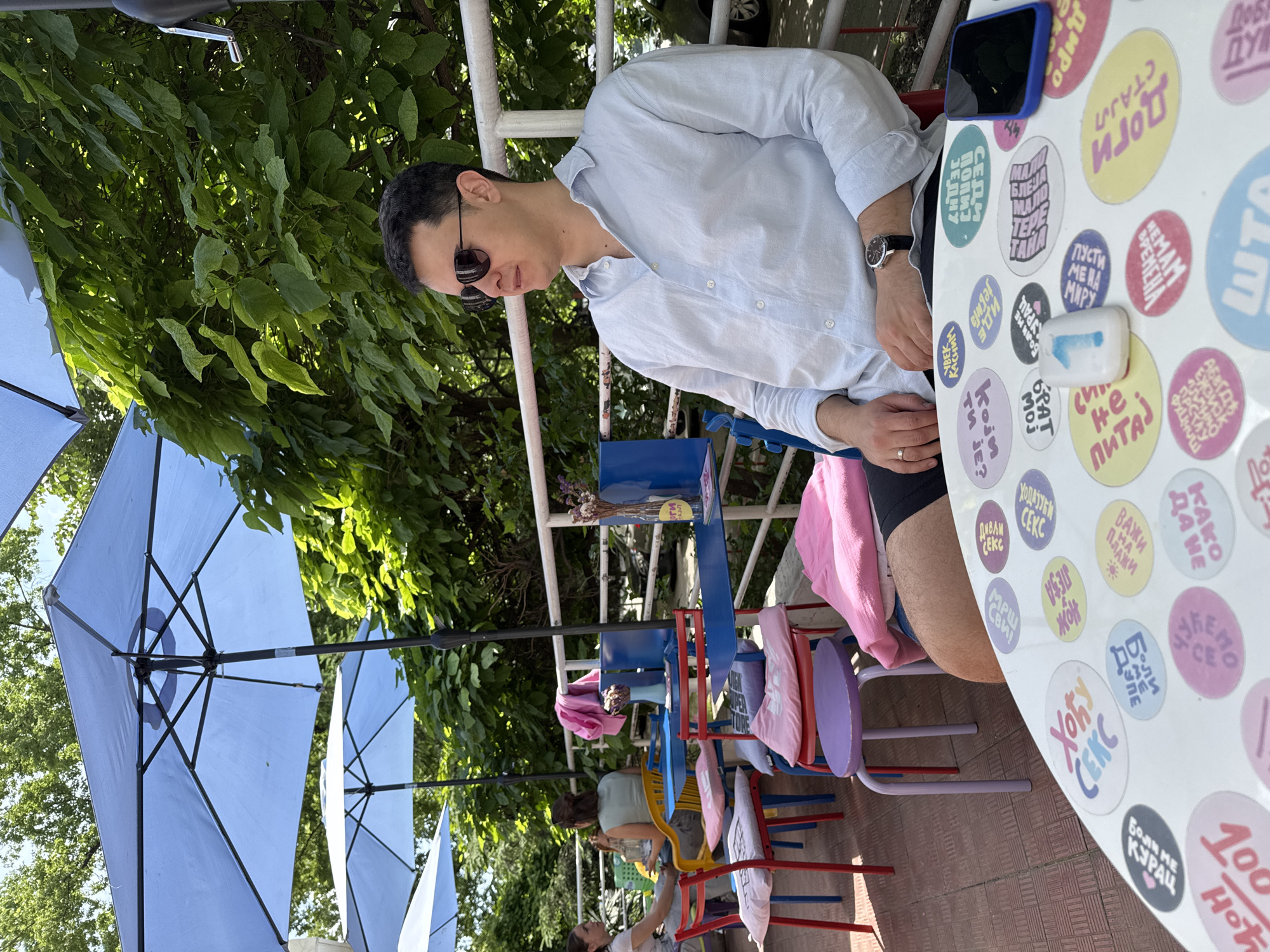 The image depicts an outdoor cafe setting, possibly in Novi Sad, Serbia, given the language on the sticker-covered table. A man wearing sunglasses and a light-colored shirt sits at a table with a phone, a small white container, and a watch visible. The table is decorated with numerous colorful stickers featuring Serbian phrases. The cafe setting includes multi-colored metal chairs and blue parasols shading the area. In the background, other patrons can be seen sitting at tables, and there is greenery suggesting a park-like environment. A metal railing partially obscures the view of the background foliage. The photo appears to be taken during daylight hours.