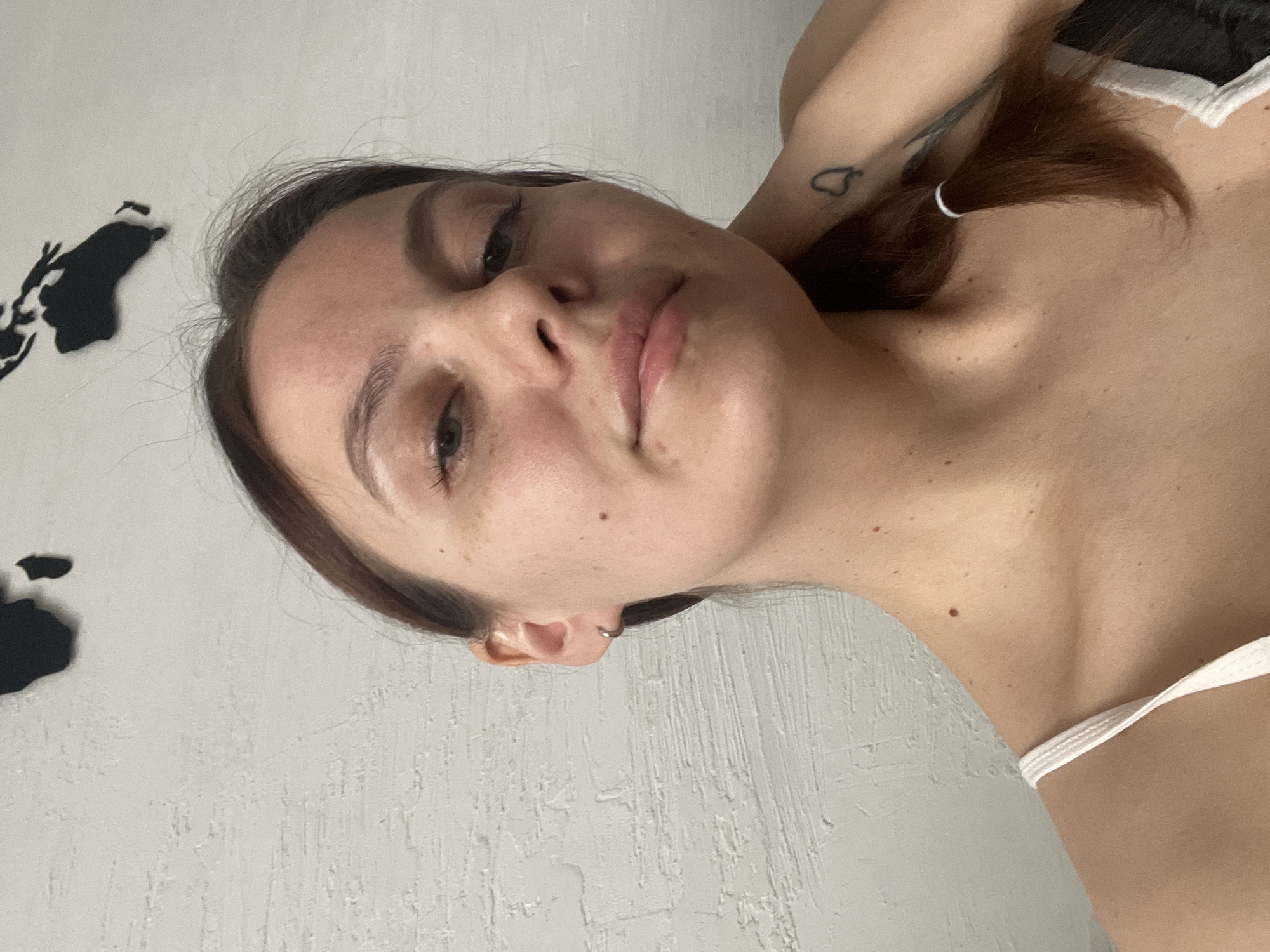 A young woman is captured in a close-up, chest-up self-portrait indoors in Chaikovsky, Russia. She has dark brown hair and a relaxed, neutral expression, looking directly or slightly past the camera. Her head is tilted, and her left arm is raised, possibly with her hand behind her head or neck. A small, black heart-shaped tattoo is visible on her upper left arm. She is wearing a white-strapped garment, possibly a tank top or bra. The background consists of a light-colored, textured wall. On the upper left side of the wall, there is a black silhouette, appearing to be a stylized map or abstract art, potentially part of a larger world map decor. The lighting is soft and diffused, indicating an indoor setting, likely during daylight hours. No discernible text is present in the image.