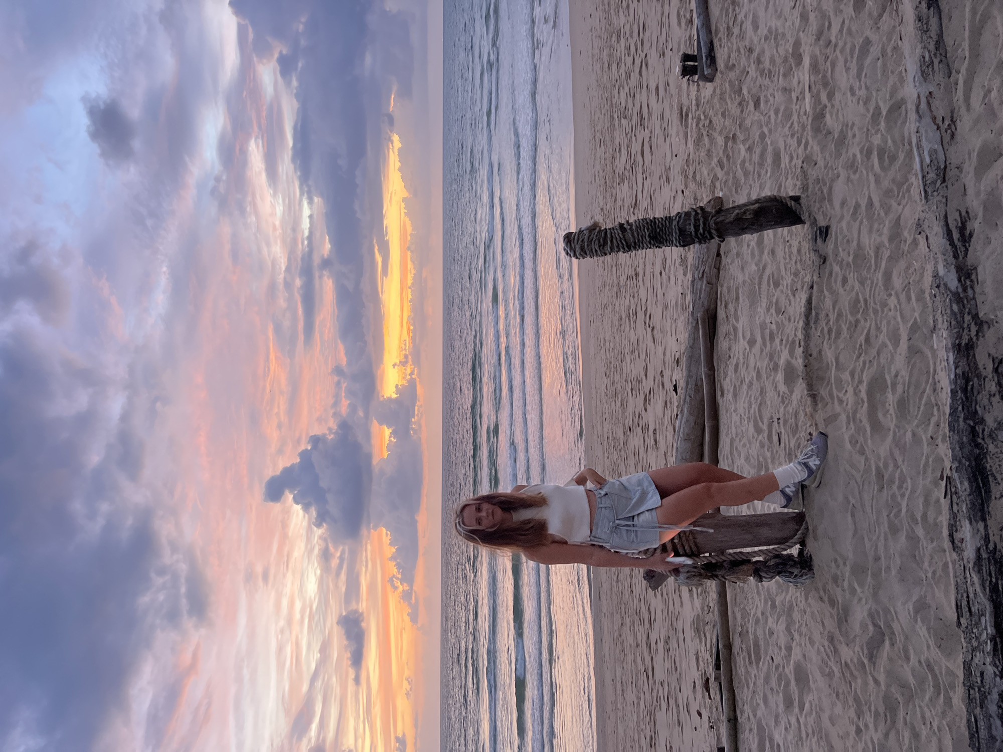 A young woman with blonde hair stands on a sandy beach in Choeng Thale, Thailand, at sunset. She is wearing a white crop top and a light blue denim skirt, with white socks and grey sneakers. Her left hand is in her skirt pocket, and she is leaning against a wooden post wrapped with rope. The ocean stretches behind her, with gentle waves and a colorful sunset sky displaying hues of pink, orange, and purple. The beach is scattered with driftwood and sand.