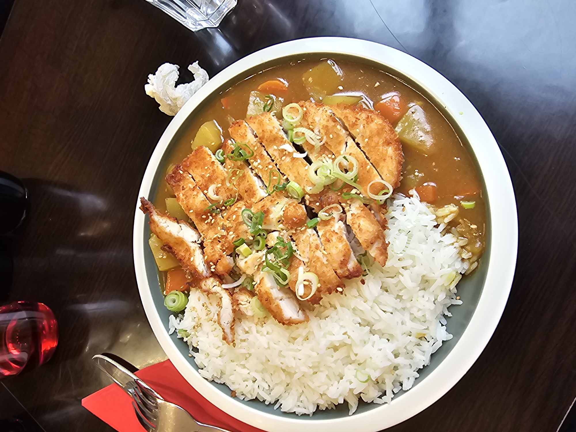 A top-down view shows a plate of Japanese chicken katsu curry on a dark wood table. The plate contains a mound of white rice, with sliced breaded and fried chicken arranged on top. The chicken is garnished with chopped green onions and sesame seeds. The curry surrounding the rice and chicken includes chunks of carrots and potatoes. A knife and fork rest on a red napkin to the left of the plate. A clear glass of water and a crumpled tissue are visible at the top left of the frame. A small red glass sits at the bottom left. The image location is likely a restaurant, but there are no specific details indicating the city of Paris, France.