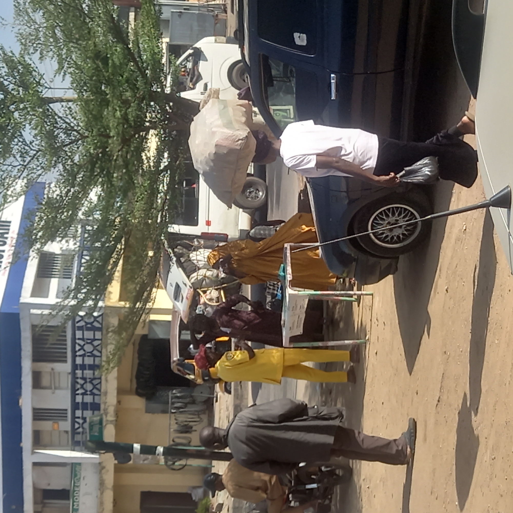 Sardauna, Nigeria. A roadside scene shows several people in a casual setting. A man is seen attempting to load a large sack onto the roof of a blue car. Next to the car, there are several individuals, one dressed in a gold outfit and another in a yellow suit. The environment includes a dirt road, a white van in the background, and a tree. Buildings and other street elements are also present, suggesting a commercial area.