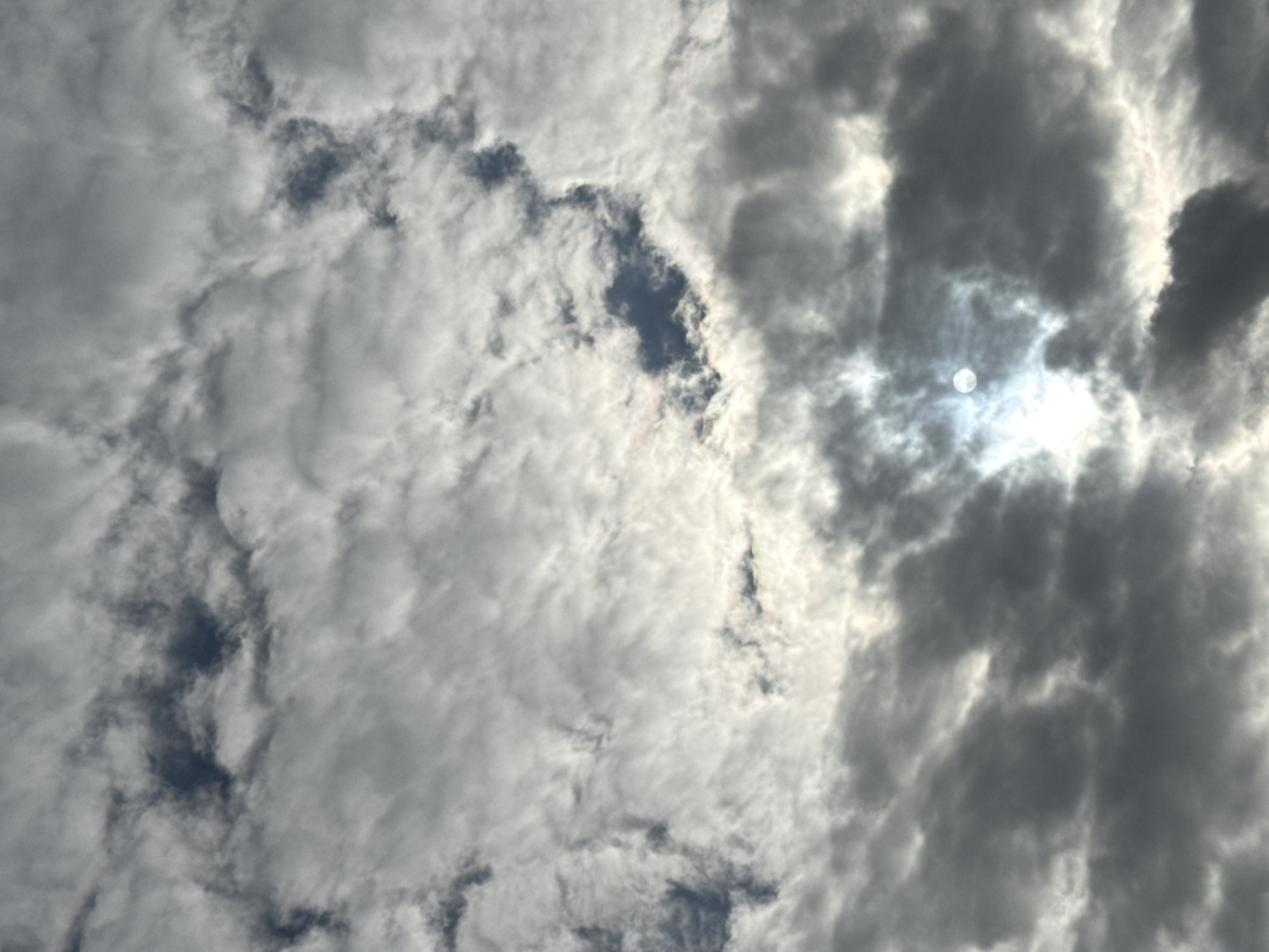 The image is an upward-facing shot of the sky from Katwijk, Netherlands, featuring a dense layer of clouds interspersed with patches of sunlight. The clouds vary in shade from light gray to dark gray, with some areas appearing almost white where the sunlight breaks through. The sun itself is partially obscured by clouds, appearing as a bright, diffused disc. The overall impression is of a partly cloudy sky with the sun attempting to penetrate the cloud cover.
