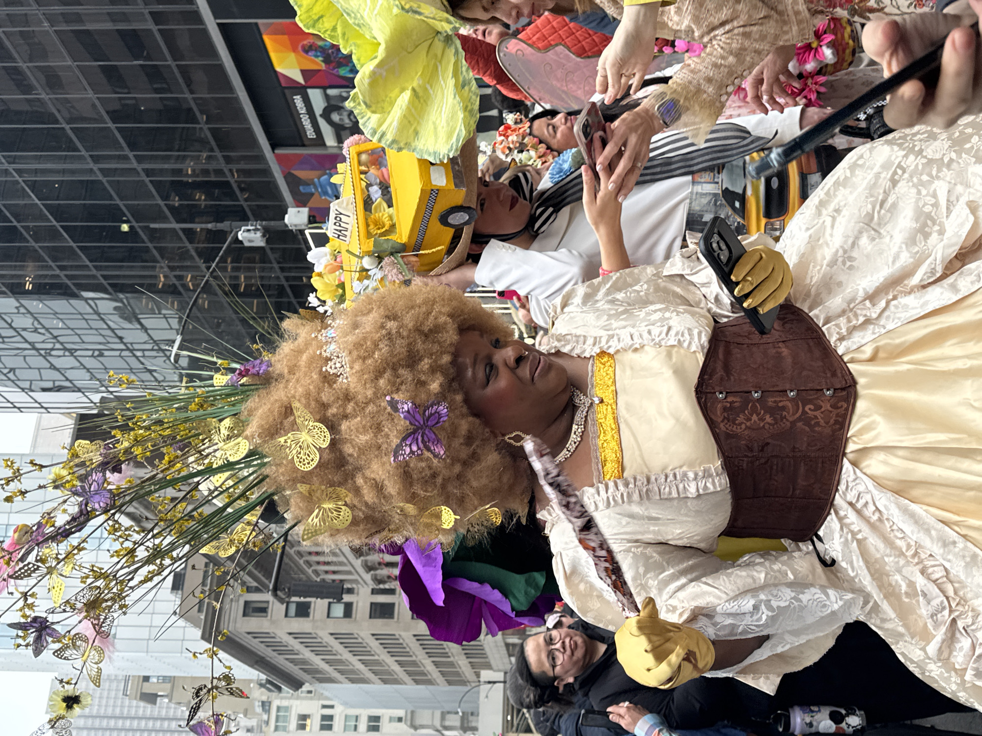 The image captures a person dressed in a fanciful, ornate costume in an urban setting, likely New York City, United States of America. The individual has a large, curly wig adorned with flowers and butterflies. They are wearing a period-style dress with ruffles, a brown corset-like garment, and yellow gloves. They are holding a phone. Several other people are around the costumed individual, some of whom are also in costume or taking photos. Buildings reflecting the sky are visible in the background.