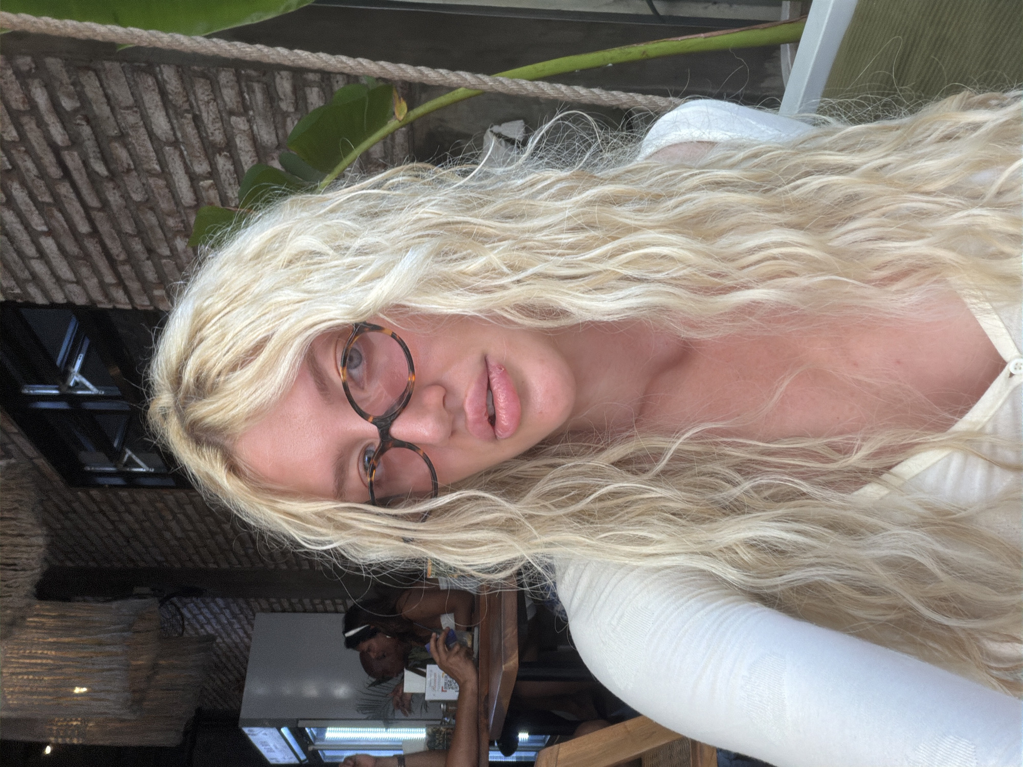 A blonde woman wearing a white top and tortoise-shell glasses is taking a selfie in what appears to be an outdoor cafe in North Kuta, Indonesia. She has long, wavy blonde hair and is looking directly at the camera. In the background, there are tables and chairs, other patrons, a brick wall, and tropical plants. The lighting suggests it is daytime.