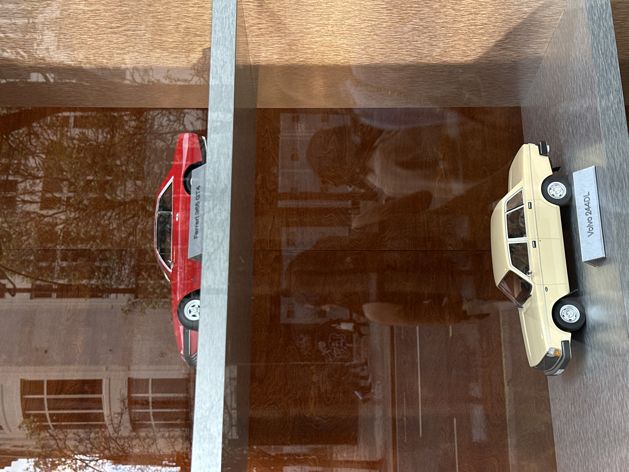 The image shows two model cars displayed in a shelf or cabinet. On the left is a red model car labeled "Ferrari 365 GT4." On the right is a cream-colored model car labeled "Volvo 244DL." The surrounding environment seems to be reflecting in the glass or surface of the shelf, revealing what appears to be a building facade, possibly in London, United Kingdom, as indicated by the style of architecture with arched windows. Trees are also visible in the reflection. The lighting appears to be natural, as evidenced by the reflections and shadows.