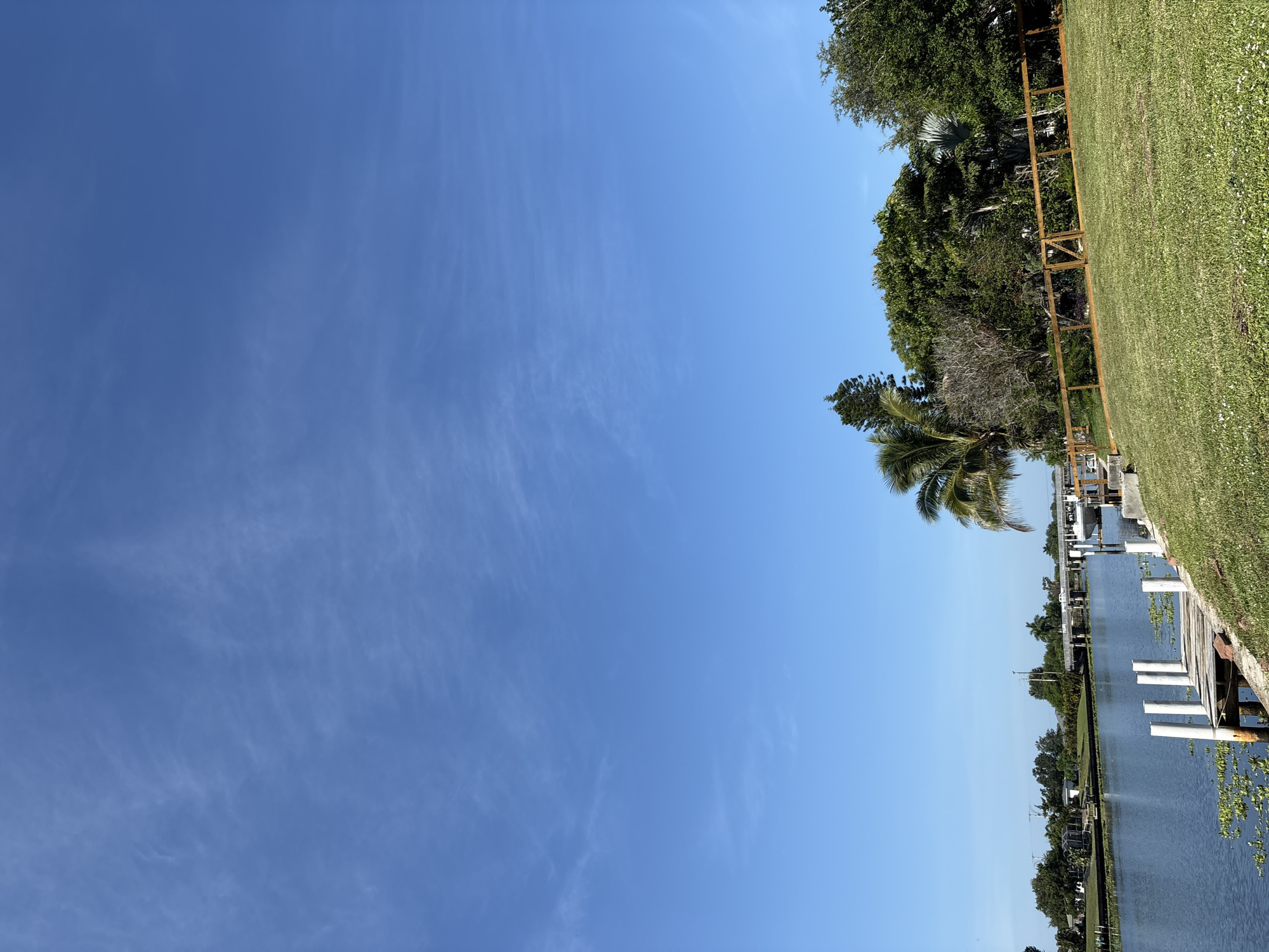 The image depicts a serene canal scene in North Port, Florida. A calm, blue canal runs horizontally across the lower left of the frame, bordered by lush green grass on the right. A wooden dock with white pilings extends into the water from the foreground. Across the canal, a residential area with houses and trees is visible, with a bridge spanning the waterway in the mid-ground. A white boat is docked on the right side of the canal. The sky is a clear, bright blue with a few wispy white clouds. The overall impression is one of peaceful, tropical living.