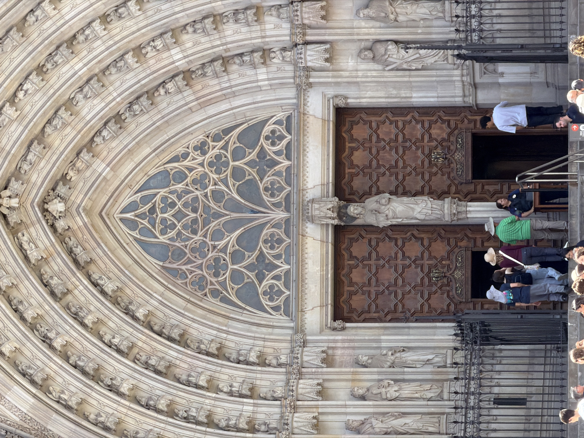 The image shows the ornate facade of the Barcelona Cathedral, a Gothic cathedral in Barcelona, Spain. The main subject is the grand entrance with its elaborately carved wooden doors and a large rose window above. Numerous statues of saints and biblical figures adorn the facade, arranged in arched niches. The architecture is detailed and weathered, indicating its age. In the foreground, a group of tourists are gathered, some looking up at the cathedral while others are interacting with a person standing near a security desk. The overall scene captures a moment of cultural tourism and admiration of historical architecture.