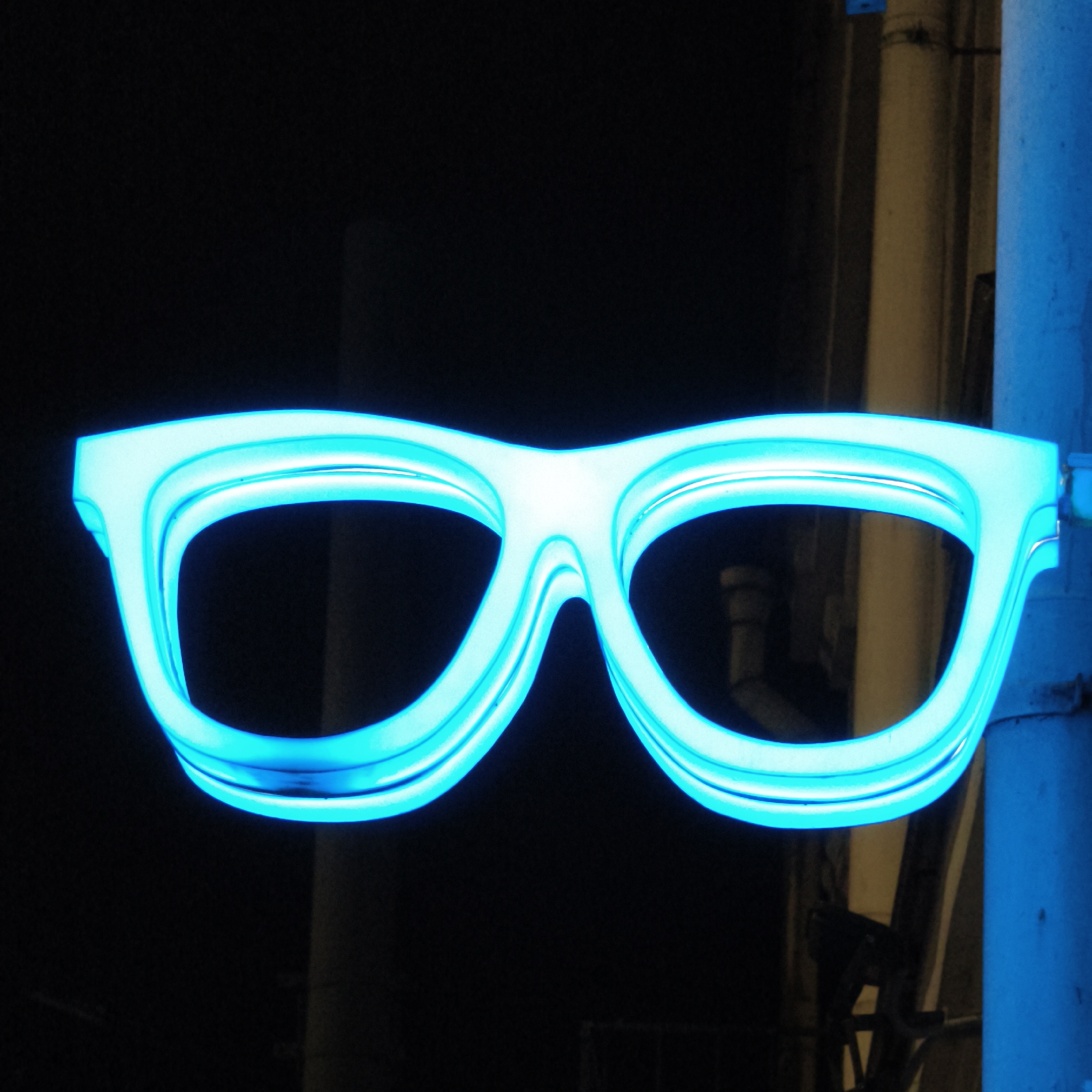 A close-up shows a glowing, neon-blue eyeglasses sign attached to a post. The background is a mix of darkness and muted colors, including a beige wall with some pipes. This sign is likely located in Hrodna, Belarus.