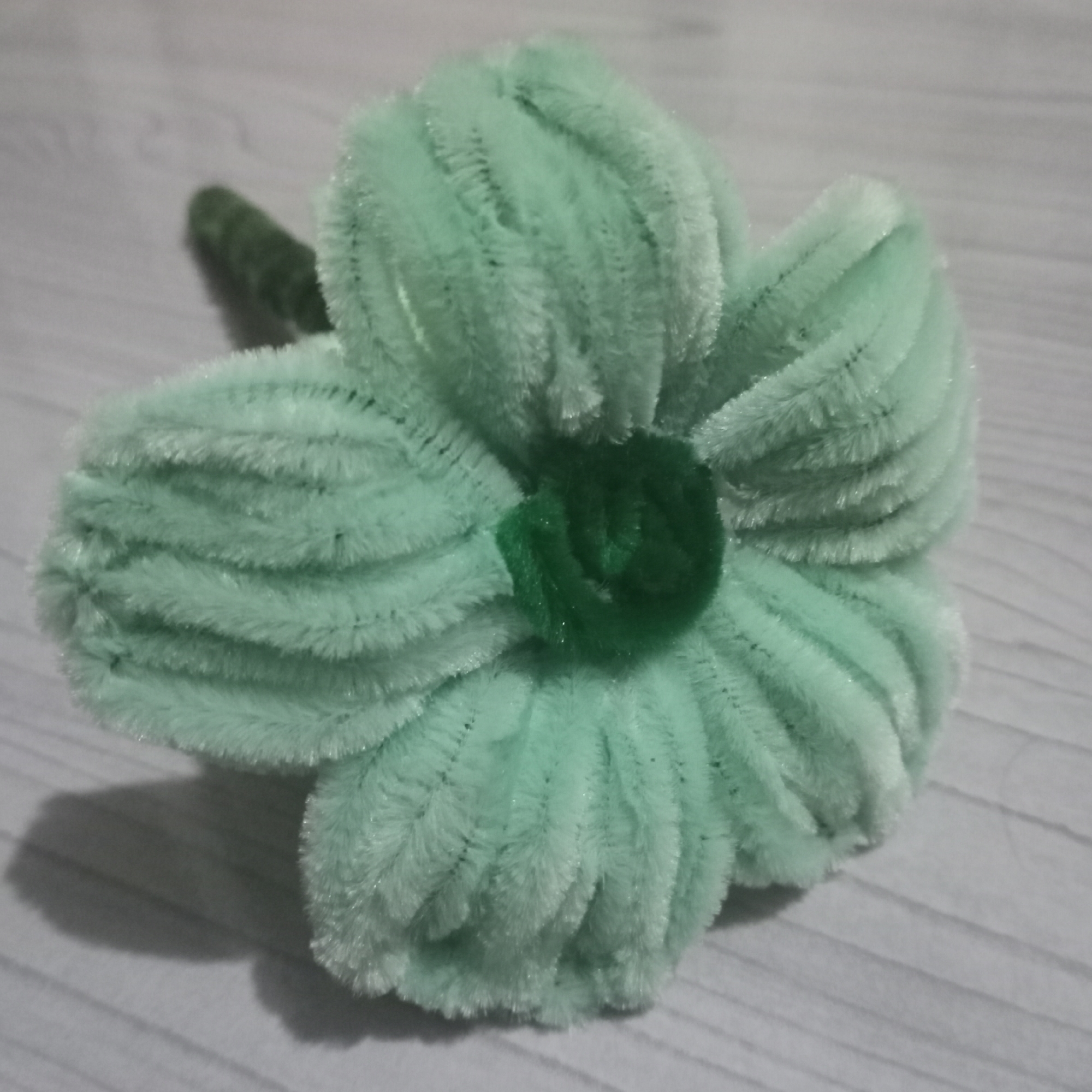 An artificial, light green, fuzzy flower made of pipe cleaners sits on a light gray surface with a wood grain texture. The flower has five petals, each with ridged detailing. In the center of the flower is a small, dark green fuzzy circle. To the left of the flower, the stem is visible, also made of green pipe cleaners. The flower is casting a shadow on the surface to its left and below. There are no people or animals present. The scene appears to be indoors, possibly on a table or desk. The location of Kedung Dowo, Indonesia is not discernible from the image.