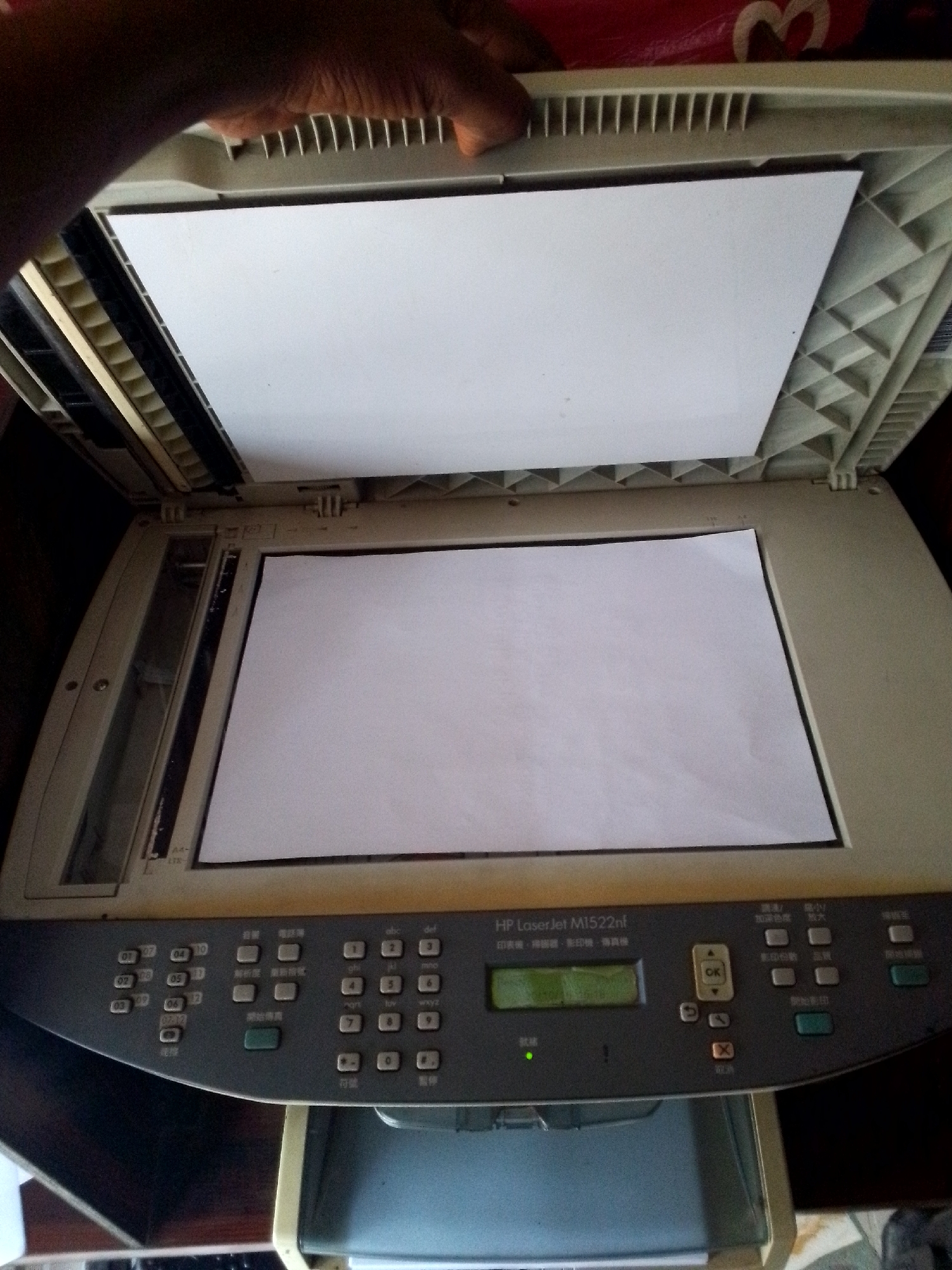 A person's hand is visible at the top, holding open the lid of an HP LaserJet M1522nf printer, which is on a table in what appears to be a home or office setting. The printer is beige and gray and has a control panel with buttons and a small LCD screen. Two sheets of white paper are visible, one resting on the scanner bed and another inside the printer's lid. The printer is not in a specific location in Nkpor, Nigeria; the image does not provide geographical context.
