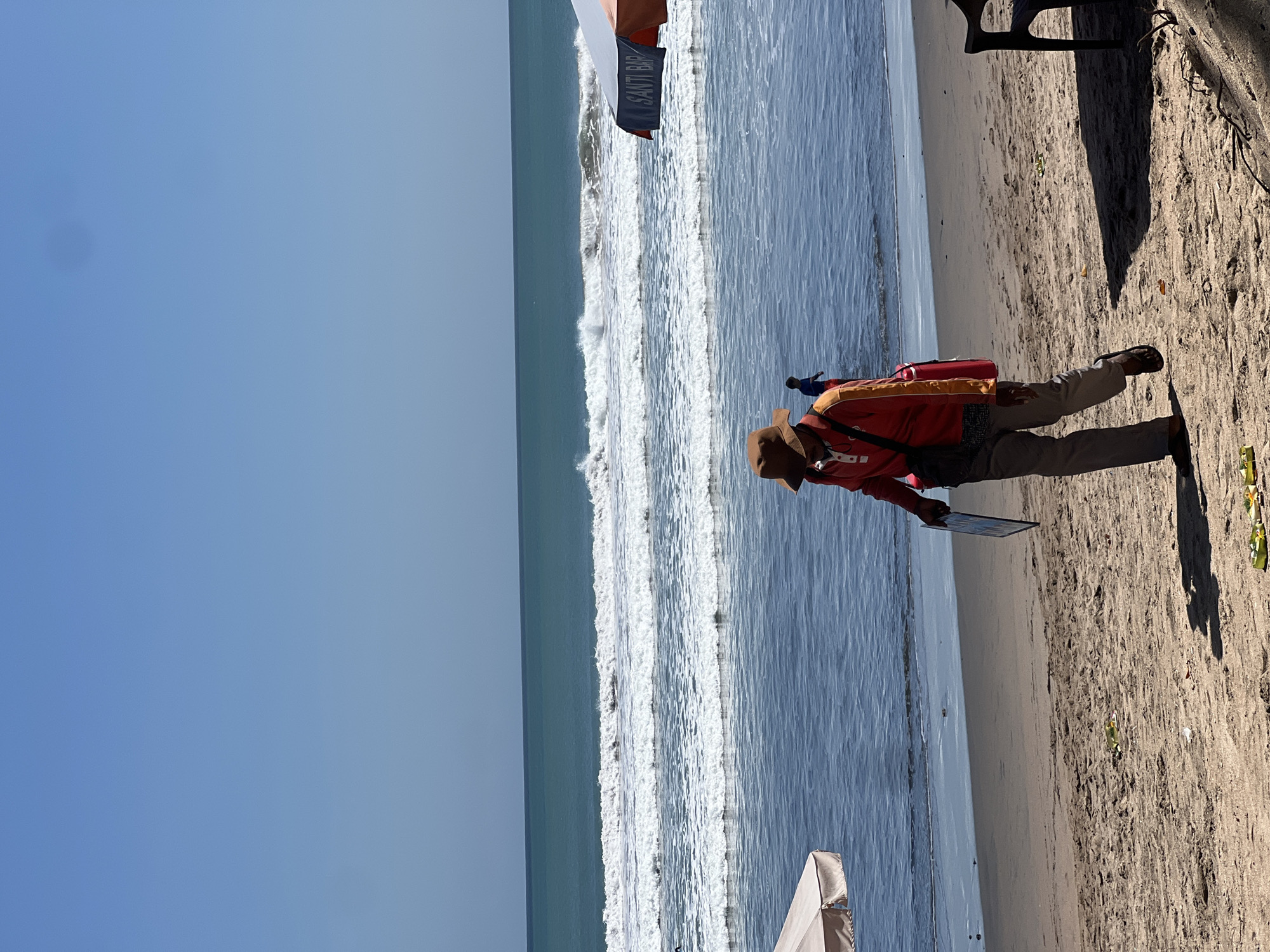 The image depicts a sunny day at Kuta Beach, Bali, Indonesia. In the foreground, a person wearing a brown hat, a red long-sleeved shirt, and grey pants walks along the sandy shore, carrying a red bag over their shoulder and a flat object in their hand. Their shadow stretches across the sand behind them. Further back, in the shallow water, another person in a blue shirt is visible. The ocean has moderate waves breaking, with white foam stretching across the horizon under a clear blue sky. An umbrella with "SANTI BAR" written on it is partially visible on the right side of the frame, suggesting a beachfront establishment. The scene is characteristic of the bustling atmosphere found at popular tourist beaches like Kuta.