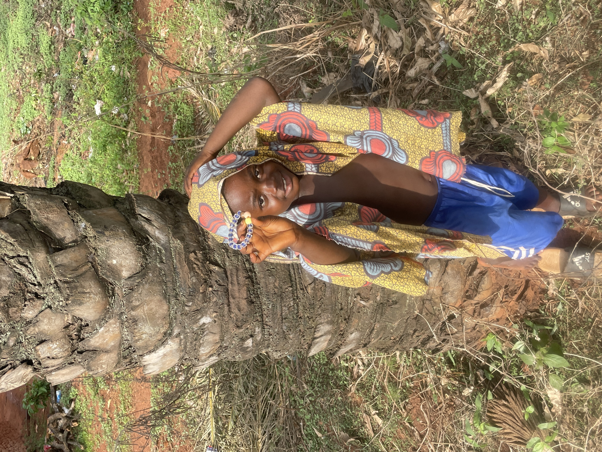 A young person is lying on a fallen palm tree trunk in an outdoor environment. The person is wearing blue shorts and a head wrap and robe featuring a yellow, red, and blue patterned fabric. They are holding a beaded bracelet in their raised hand, smiling, and looking at the camera. The palm tree trunk is brown and textured. The ground is covered with grass, leaves, and other vegetation. The location is Ogbe, Nigeria.