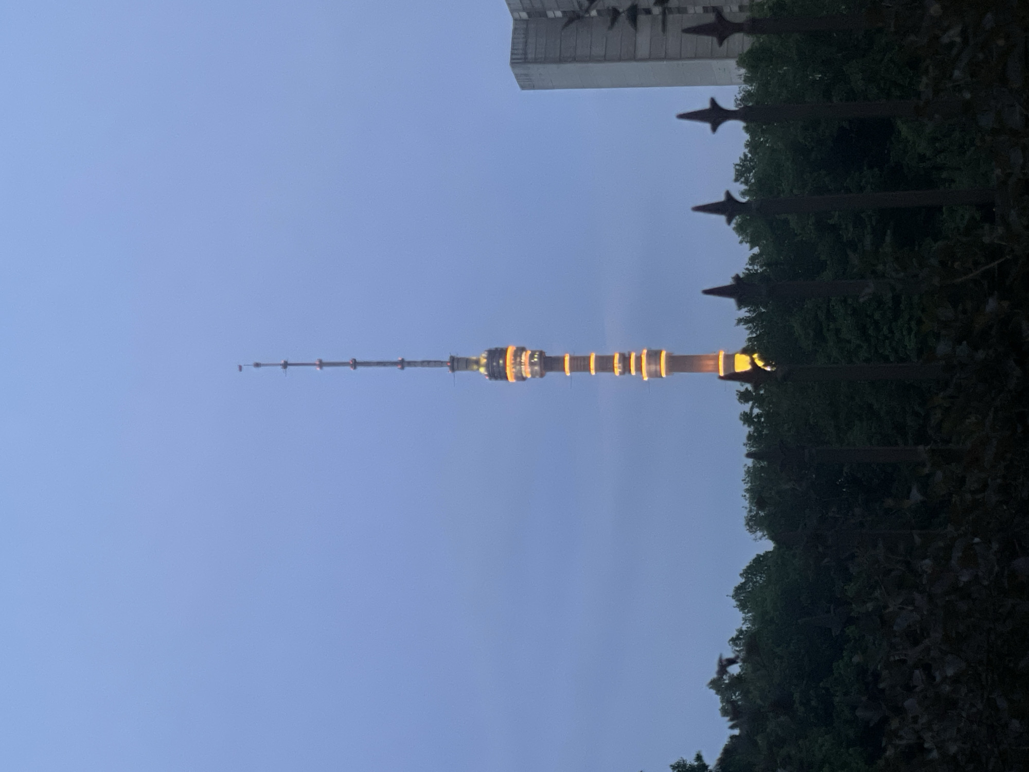 The image shows the Ostankino Tower in Moscow, Russia, at dusk. The tower is illuminated with yellow lights. The sky is a gradient of light blue to darker blue. A wrought iron fence and trees are visible in the foreground, with a building in the background. The image is taken from a low angle, looking up at the tower.