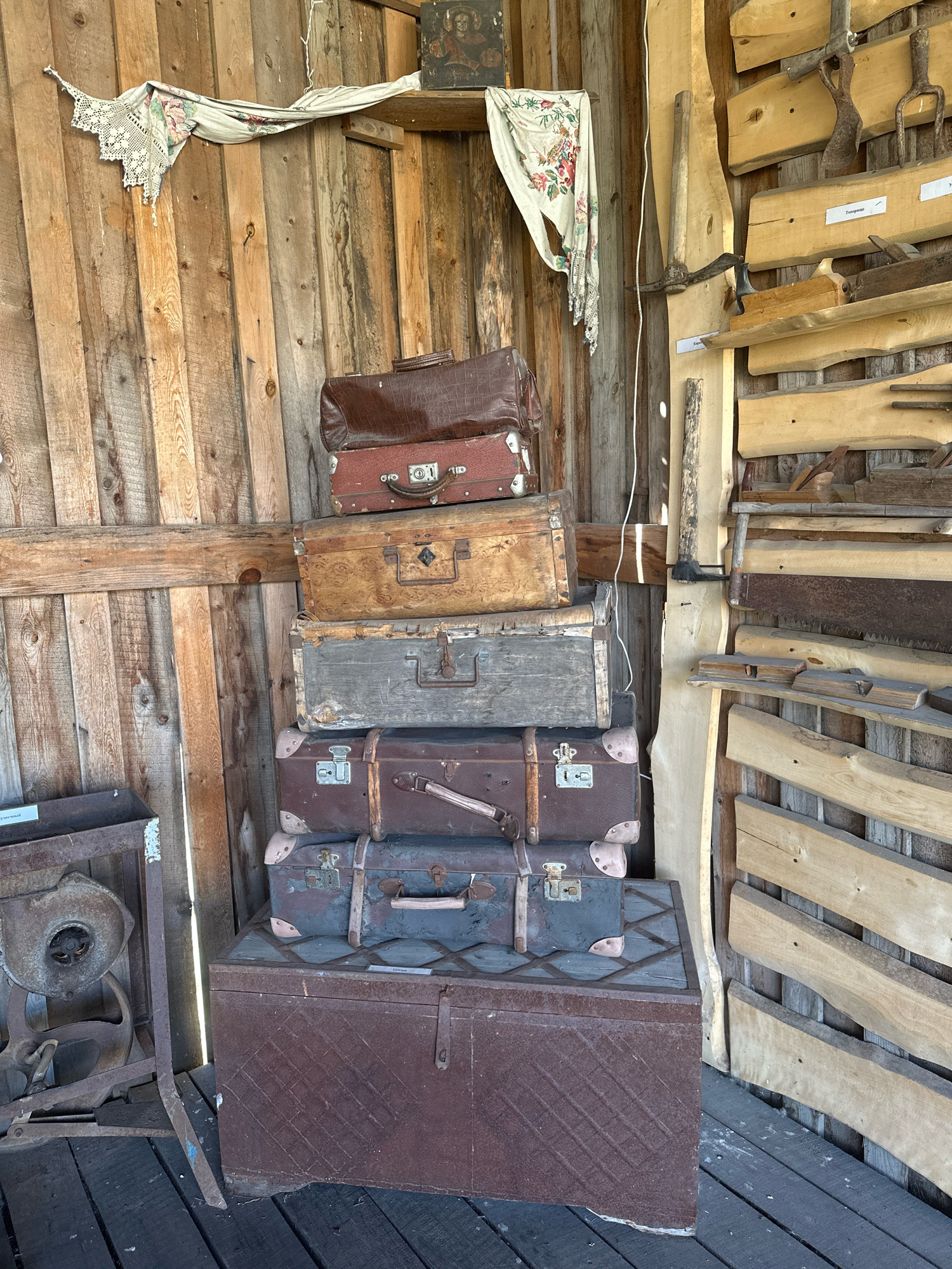 In a rustic wooden shed, a collection of vintage luggage is stacked, including a large, dark brown trunk at the base, topped by several suitcases of varying sizes and materials, the uppermost being a dark red, crocodile-textured one. The shed's interior is lined with weathered wooden planks. To the right, a wall displays an assortment of old tools, including saws, axes, and woodworking planes, mounted on wooden shelves. A piece of embroidered fabric with floral patterns hangs from a hook on the wall. In the lower left corner, a rusty piece of machinery, possibly a blower or fan, is partially visible. The floor is made of dark wooden planks. The scene appears to be in an outdoor or semi-outdoor storage area, likely a shed or barn, in the city of Kargat, Russia. There are no people or animals visible in the image.