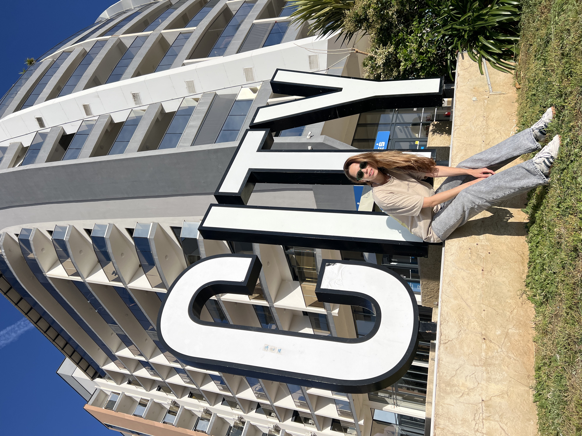 A woman is sitting on a concrete ledge in front of a large white sign that reads "CITY" in black outline letters. The building behind the sign is a modern, multi-story structure with a rounded facade and numerous windows. The background includes some greenery and a clear blue sky. The woman is wearing sunglasses, a t-shirt, and jeans. The setting appears to be an outdoor public area, likely in Bar, Montenegro, due to the presence of the "CITY" sign often associated with a hotel or attraction in that location.
