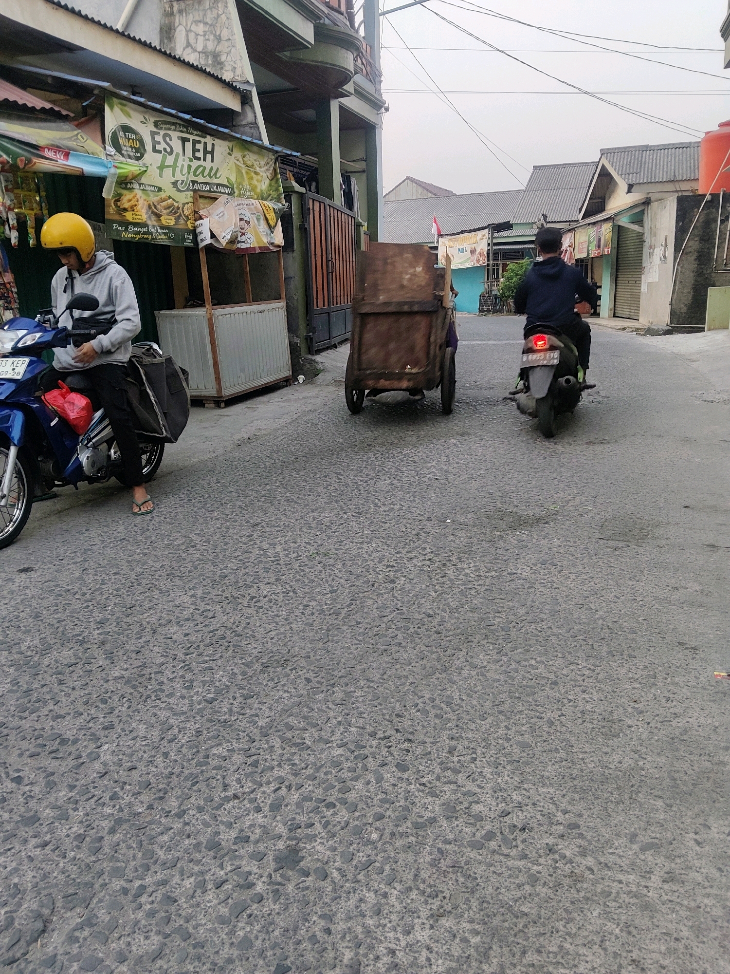 In Mekarmukti, Indonesia, a street scene unfolds with two individuals on motorcycles. On the left, a person wearing a yellow helmet and a grey hoodie sits on a blue motorcycle, facing forward. A red bag is attached to the front of the motorcycle, and a large black bag is behind the rider. The motorcycle's license plate reads "33 KEP GP". To the right, another person in a dark blue hoodie rides a dark-colored motorcycle away from the viewer. This motorcycle's taillight is illuminated, and its license plate appears to be "B 6933 FYO". Between the two motorcycles, a brown, two-wheeled cart with a wooden frame is positioned in the street. The environment features buildings with textured walls and a cloudy sky overhead. The street surface is paved with small, gray stones. A banner advertising "ES TEH HIJAU" is visible on a building to the left, along with a depiction of fried food.