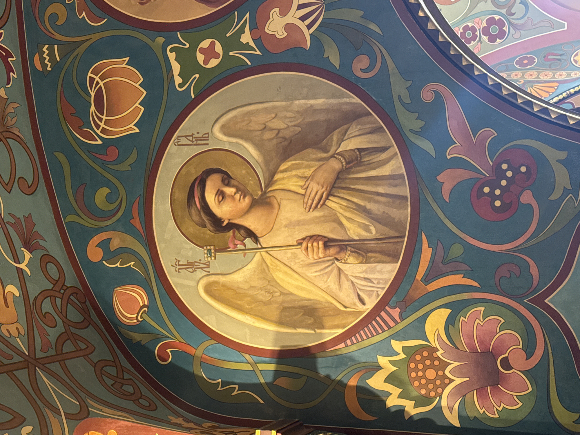 The image shows a detail of a fresco on the ceiling of the Church of the Savior on Spilled Blood in Saint Petersburg, Russia. The fresco is circular and depicts an angel with a halo, wings, and a staff. The angel is depicted from the waist up, in profile, and resting in a horizontal manner, with eyes closed. The angel is wearing a light-colored gown and holds the staff in both hands. The background of the fresco is decorated with floral and geometric patterns. The colors are mostly muted, with blues, greens, browns, and golds predominating.
